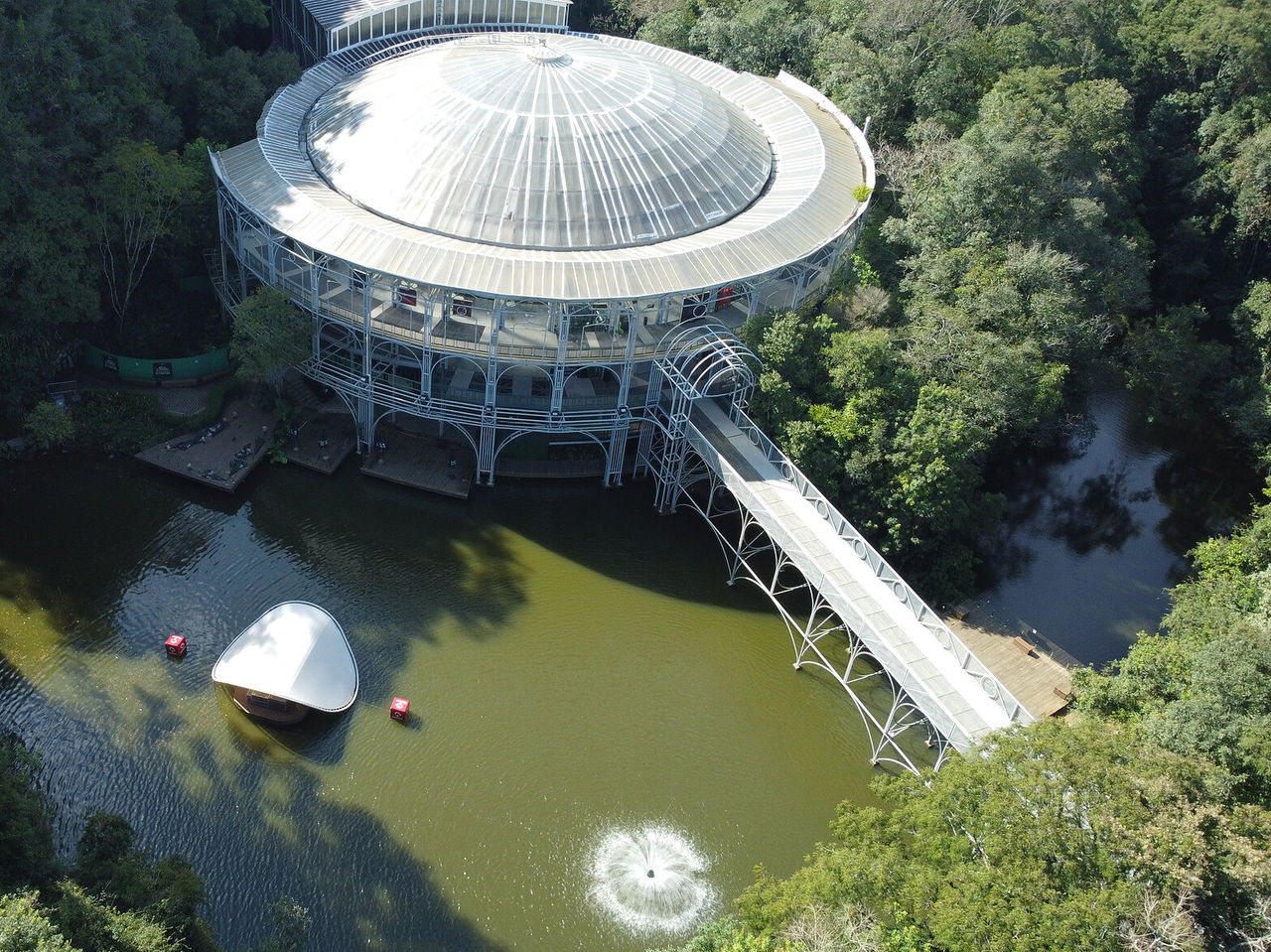 Vista aérea de la Ópera de Arame, con su estructura redonda icónica, rodeada de naturaleza, con un puente y un lago