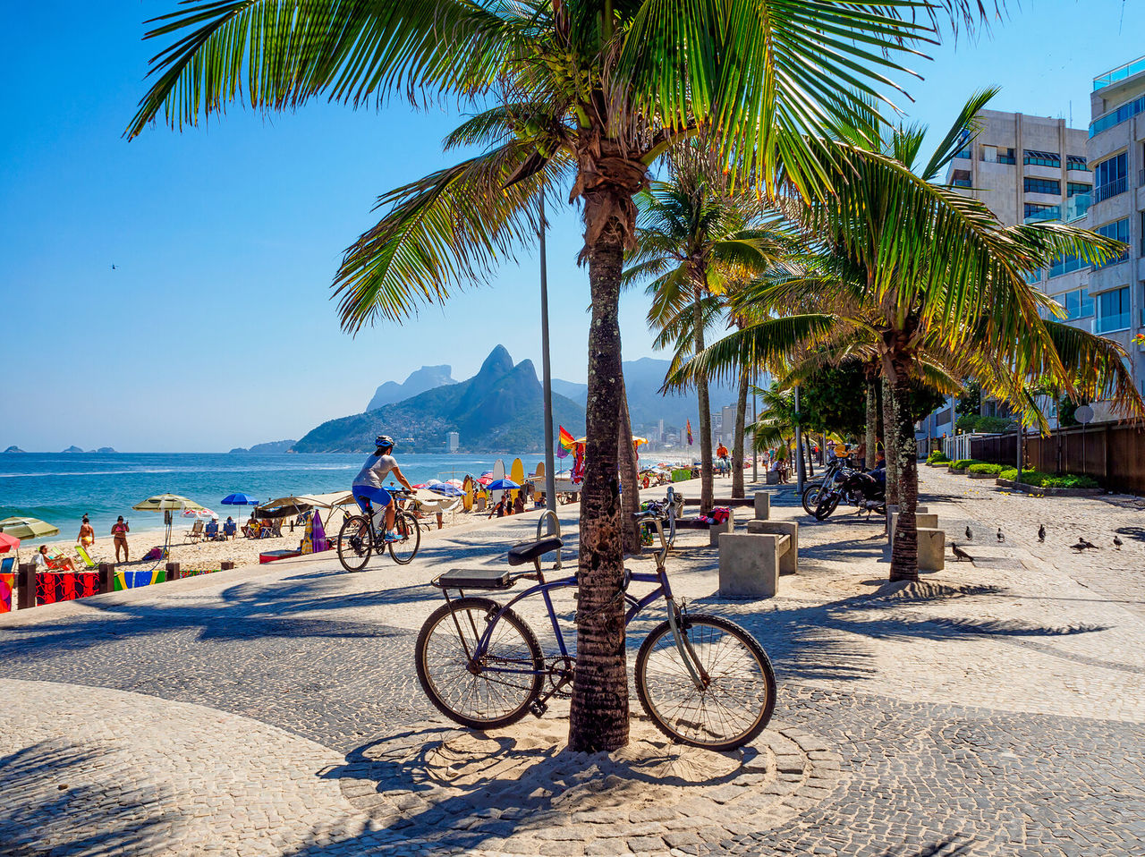 Vista de la playa de Ipanema, con personas tomando el sol, ciclistas y el morro de los Dois Irmãos de fondo