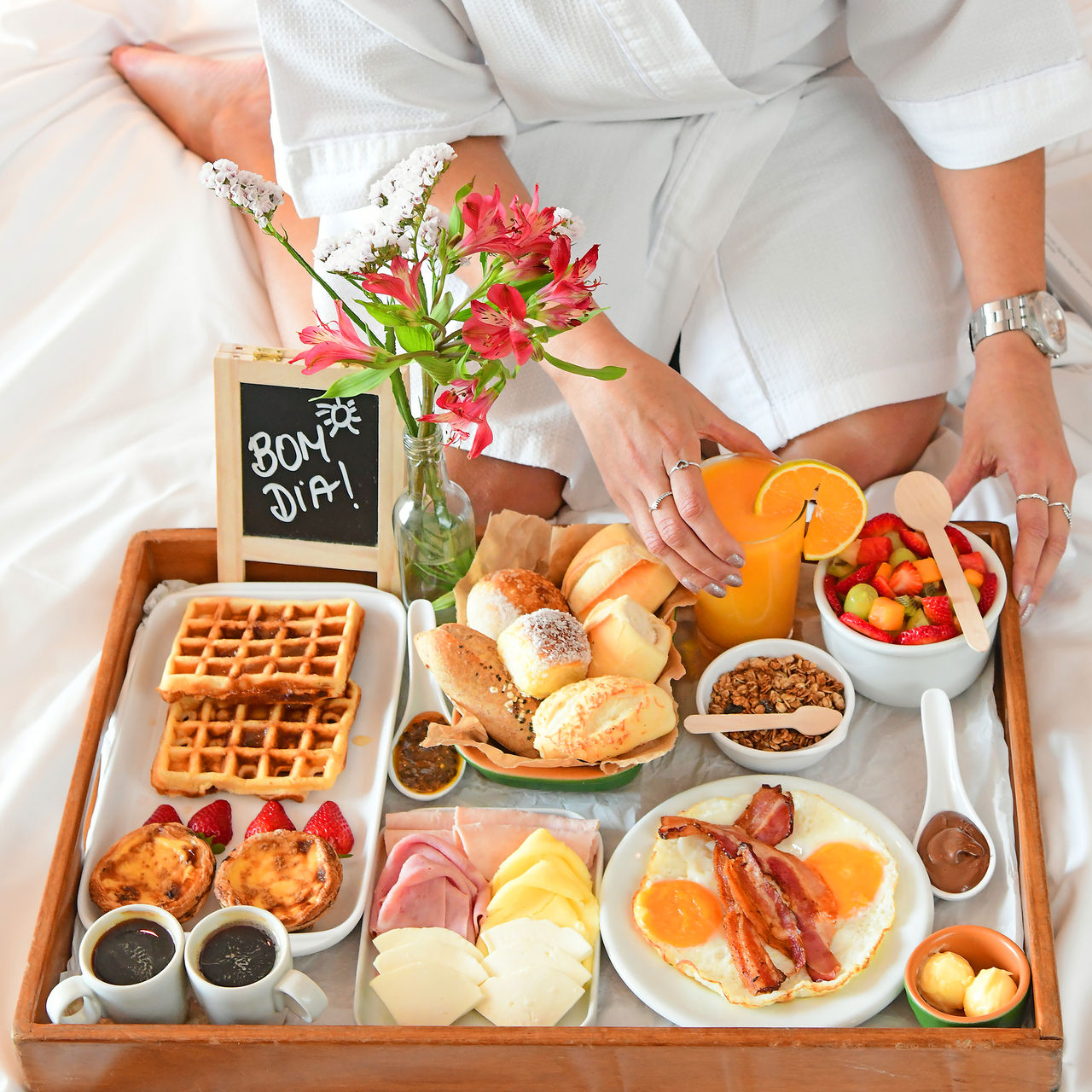 Chica en albornoz tomando el desayuno en la cama, una bandeja con pan, zumo, café, waffles, queso, jamón, fruta y huevos
