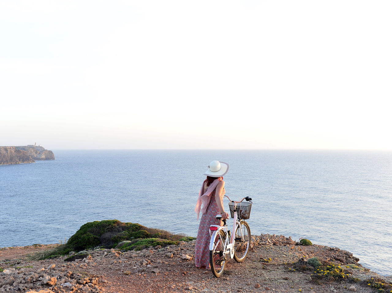 Mujer joven con sombrero y bicicleta en mano, disfrutando de la vista al mar desde un acantilado en el Algarve