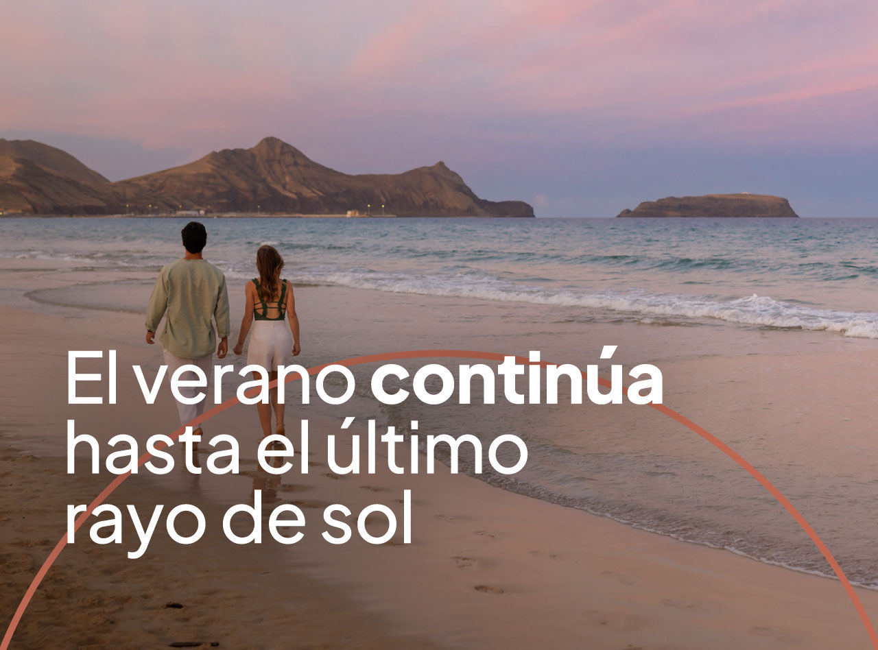 Pareja caminando por la orilla al atardecer, con montañas áridas y el mar tranquilo bajo un cielo rosado