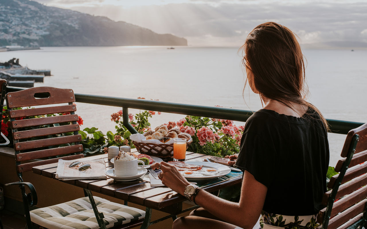 Pessoa tomando café da manhã em varanda com flores e vista para o mar iluminado entre montanhas ao fundo