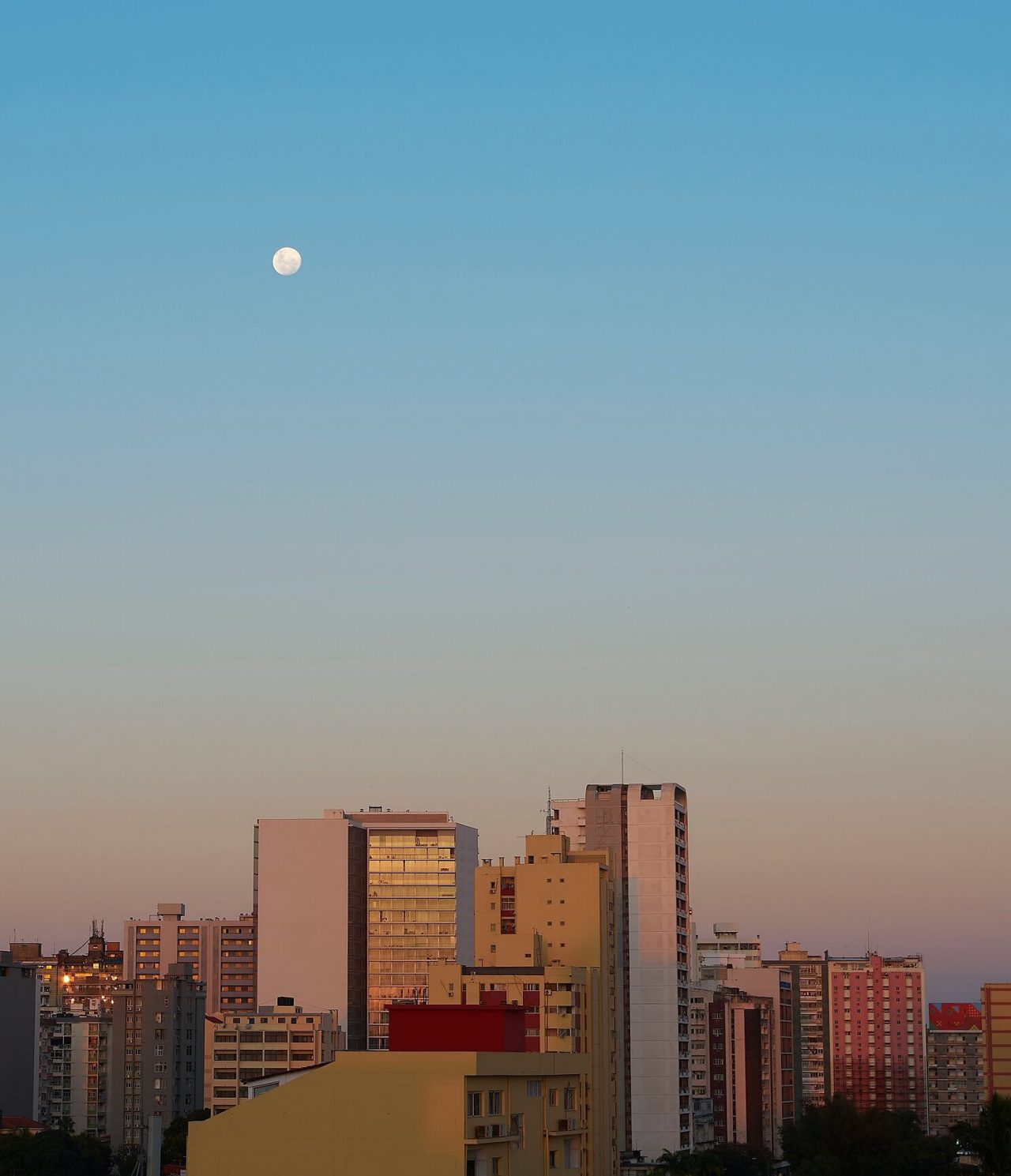 Vista da parte de cima de vários prédios de Maputo, com a luz de lusco-fusco a incidir sobre eles e a lua já alta no céu