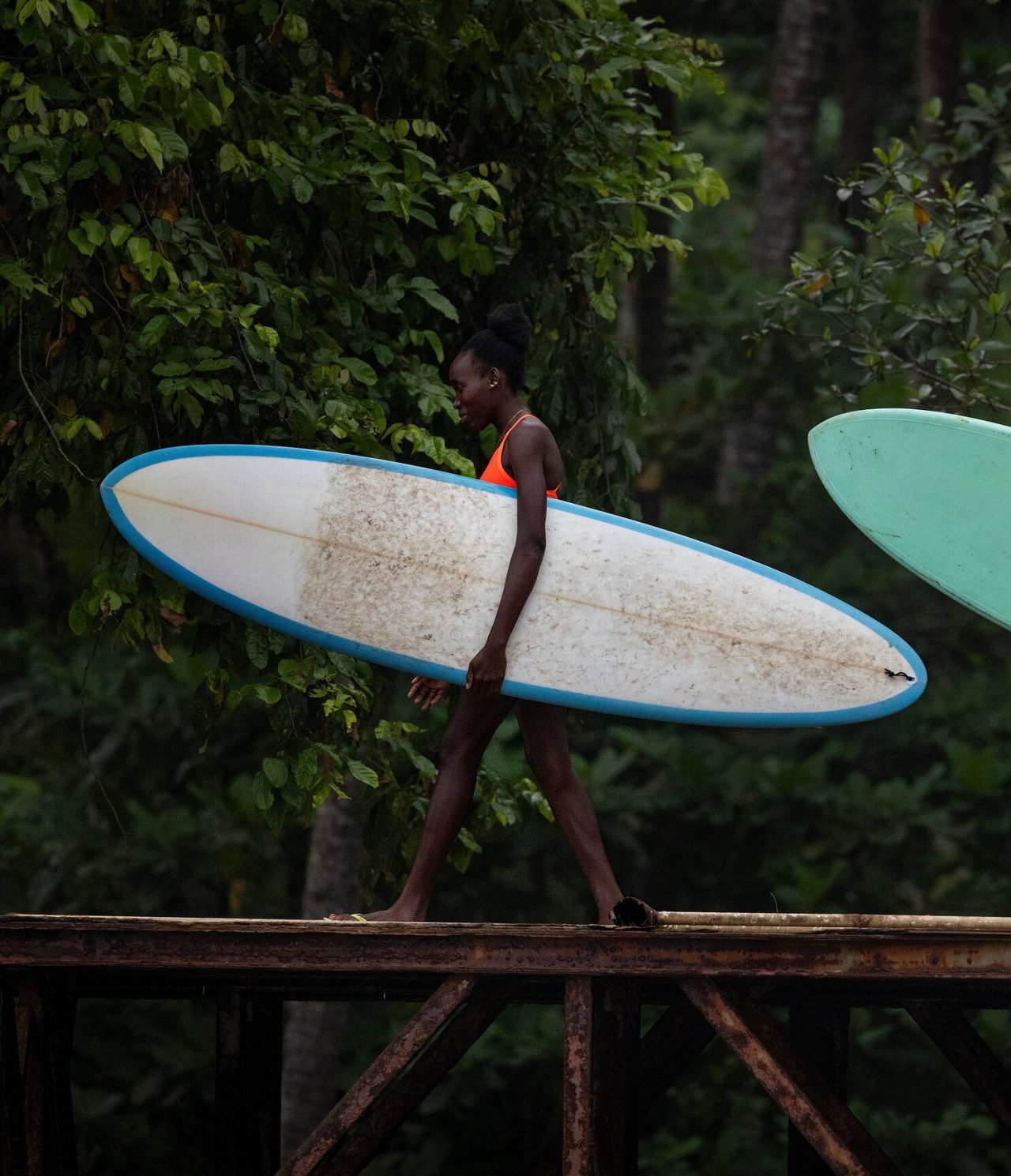 Mulher em São Tomé a andar sobre uma ponte de ferro, com uma prancha de surf na mão, com vegetação atrás