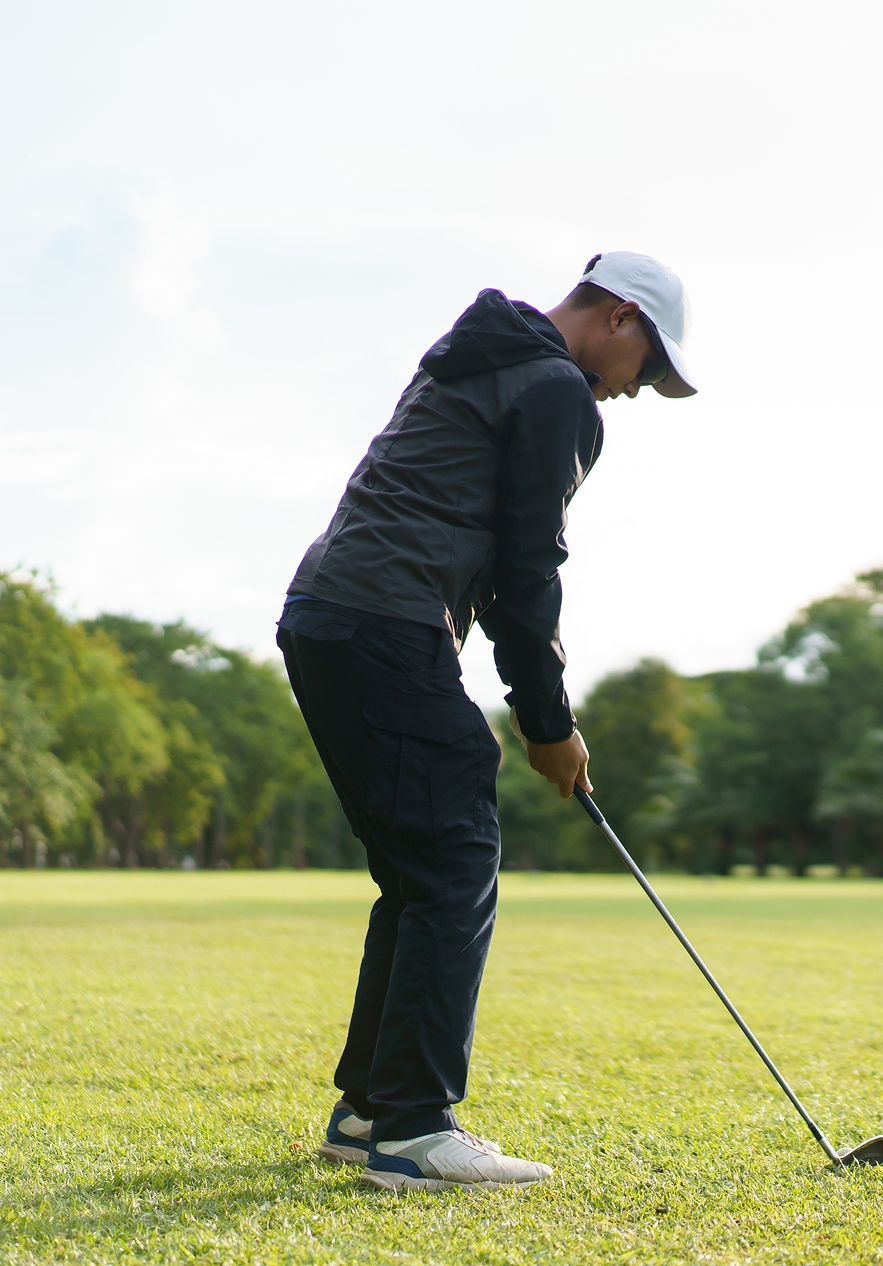 Homem de chapéu branco e roupas desportivas a jogar golfe num campo muito verde com árvores desfocadas ao longe