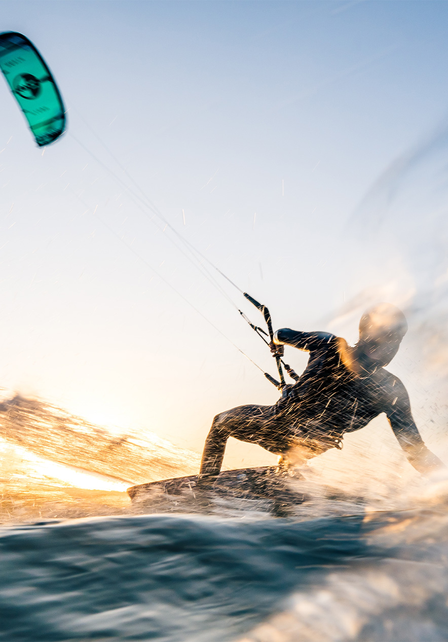 Senhor a fazer kite-surfing no mar com a vela azul e água a salpicar, com o sol a refletir na água