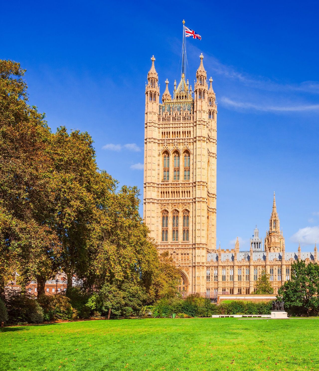 Vista de um jardim com um palácio, com uma arquitetura típica de Londres, com árvores à frente e a bandeira no topo