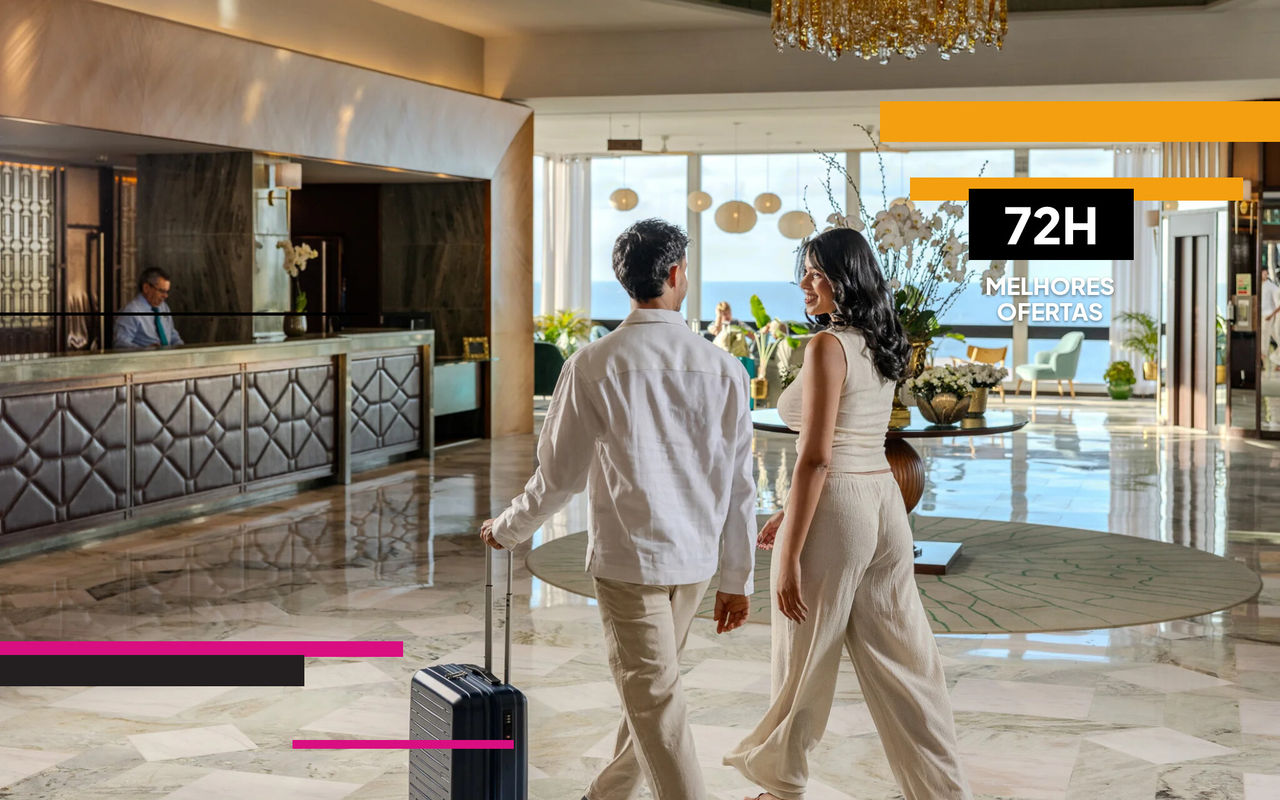 Casal com malas caminha pelo lobby iluminado de hotel com recepção ampla, lustre dourado e vista ao mar