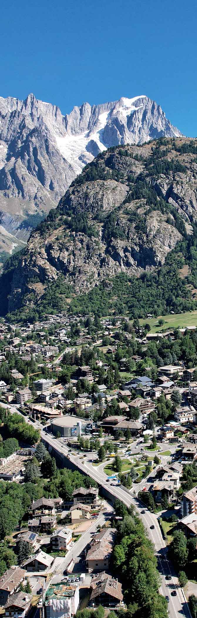 Luftaufnahme eines alpinen Dorfes mit Straßen und Häusern vor schroffen schneebedeckten Bergen unter blauem Himmel