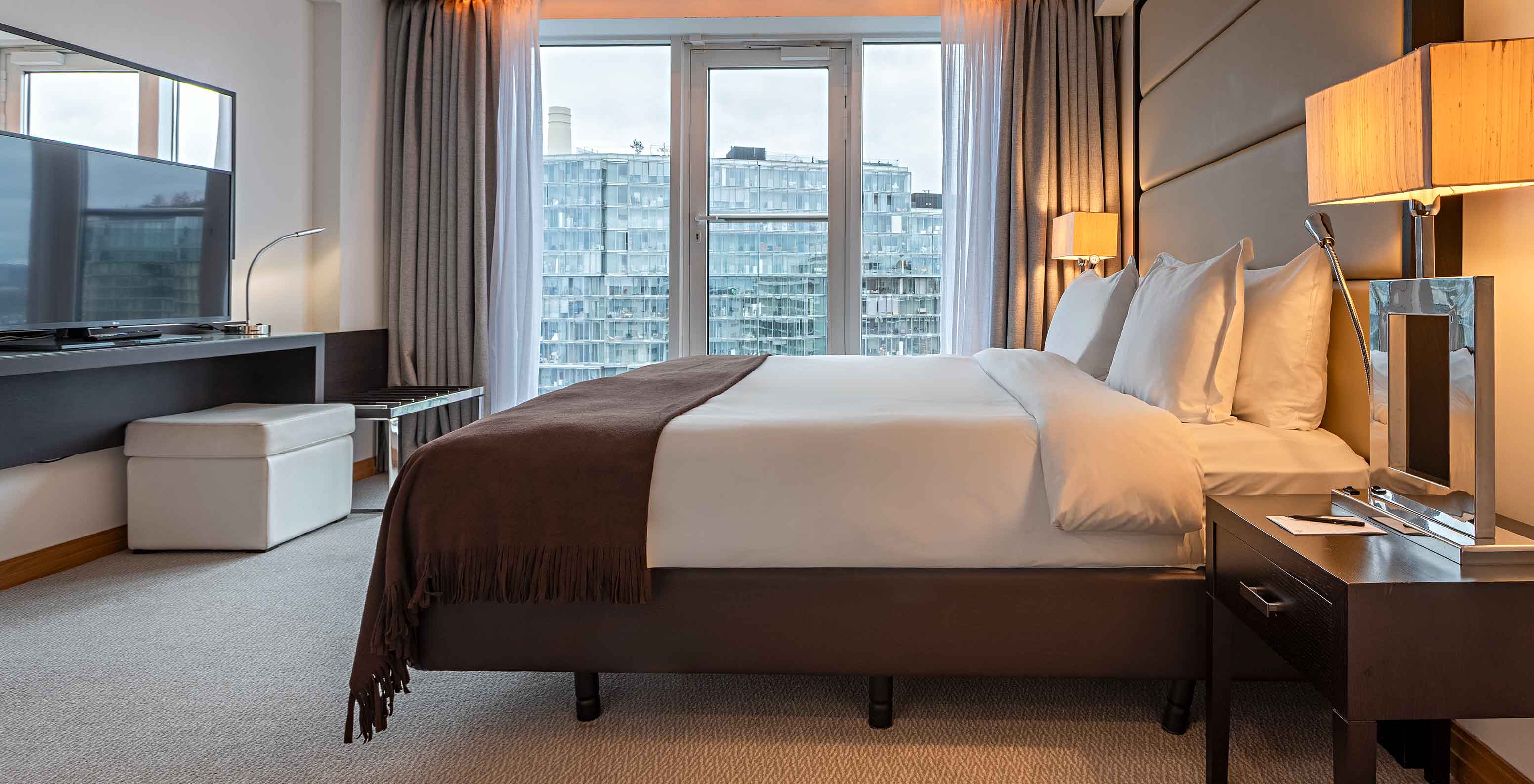 Modernes Schlafzimmer mit großem Bett, Nachttischen, Fernseher und Fensterblick auf Wohngebäude im Tageslicht