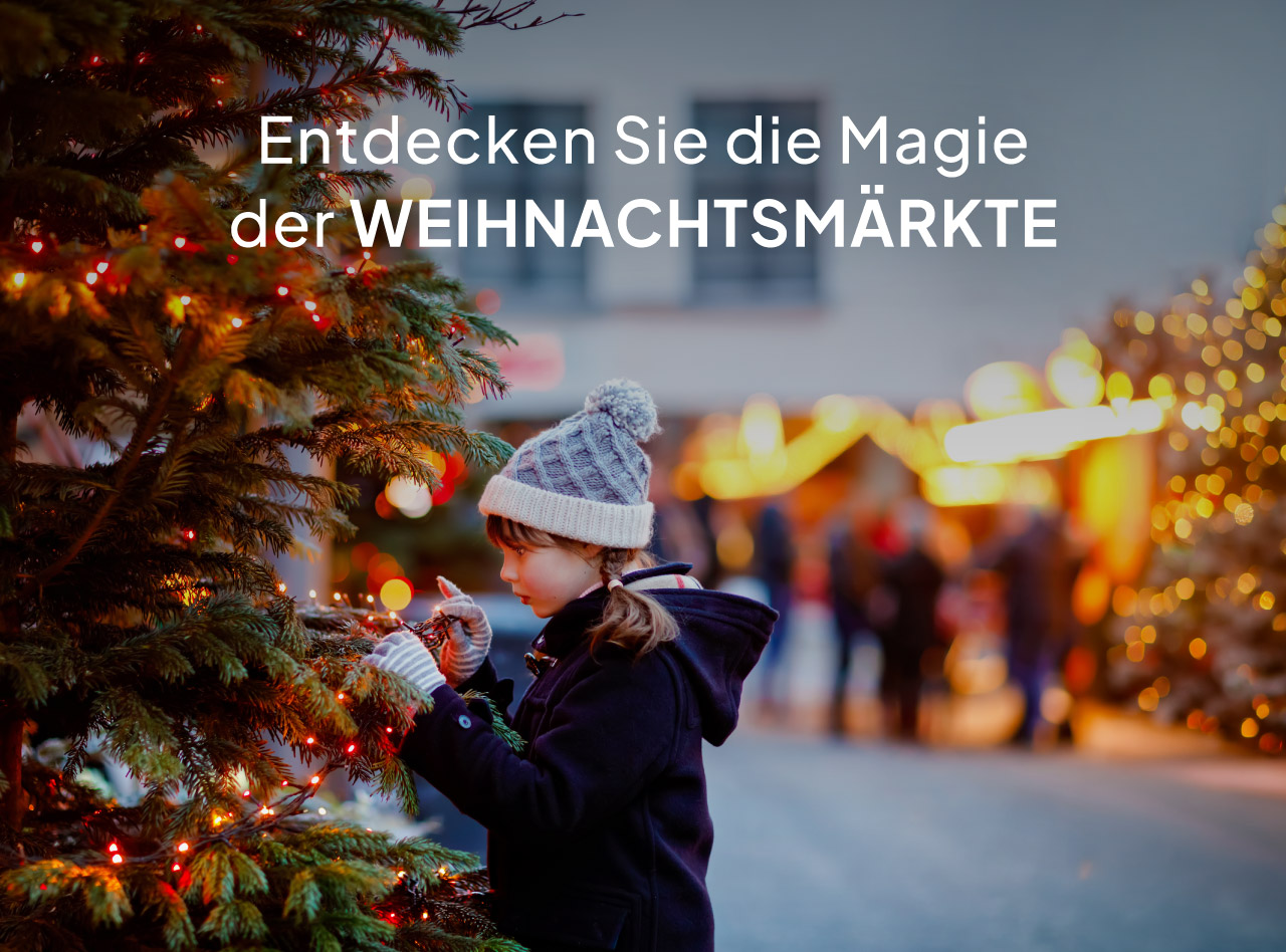 Kind mit Wintermütze schmückt einen beleuchteten Weihnachtsbaum vor unscharfen Marktständen am Abend