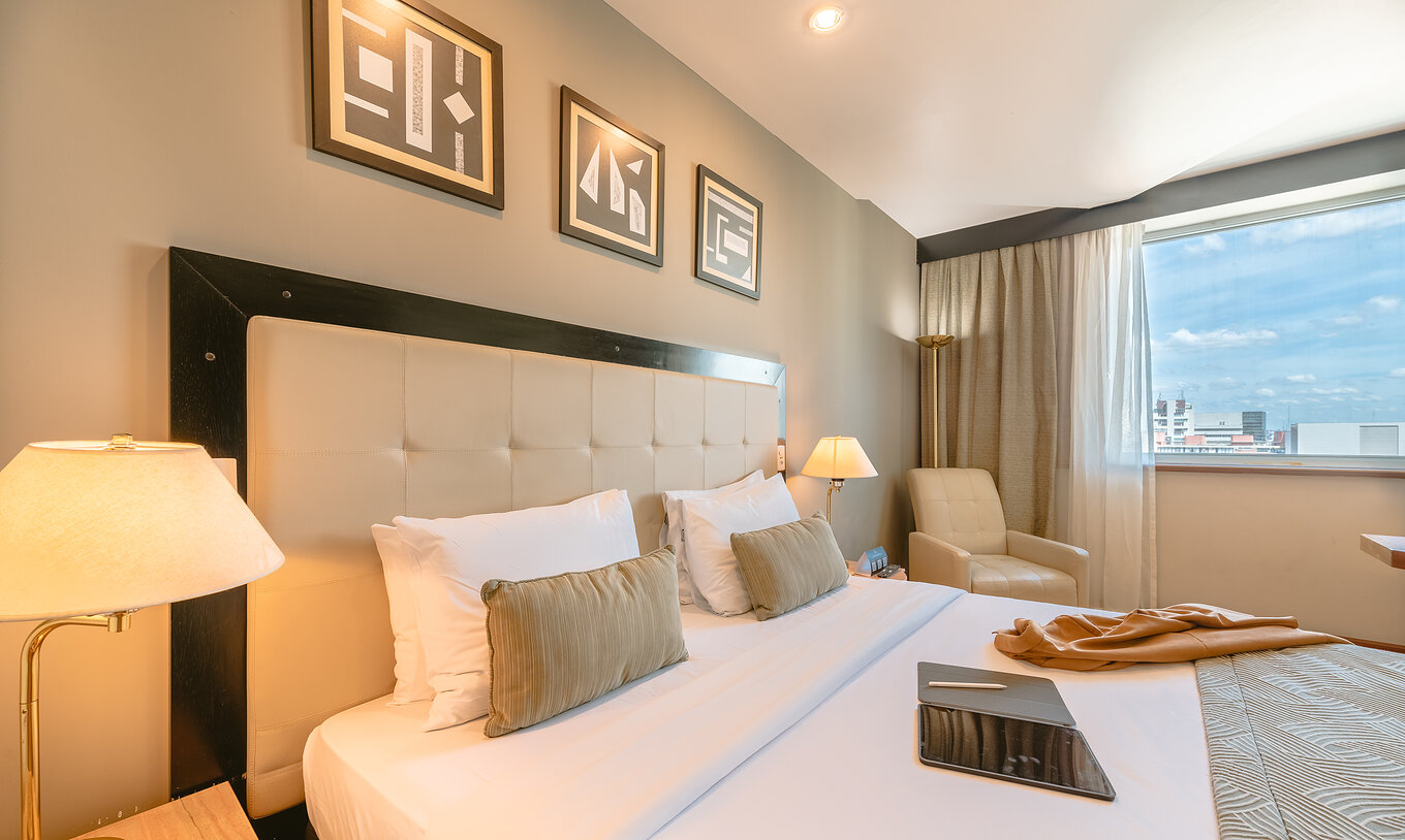 The Queen Standard of Pestana São Paulo has a double bed with a beige headboard and a window with city view
