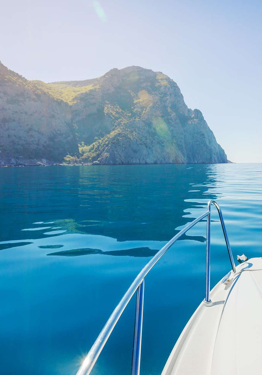 Boat gliding across calm blue water beside steep rocky cliffs under bright sunlight on a clear coastal day