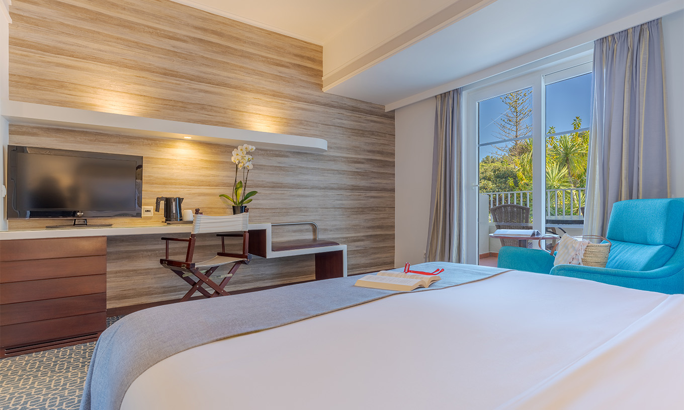 The Deluxe at Pestana Alvor Praia has a double bed, a desk TV, a blue armchair, and a window to the balcony