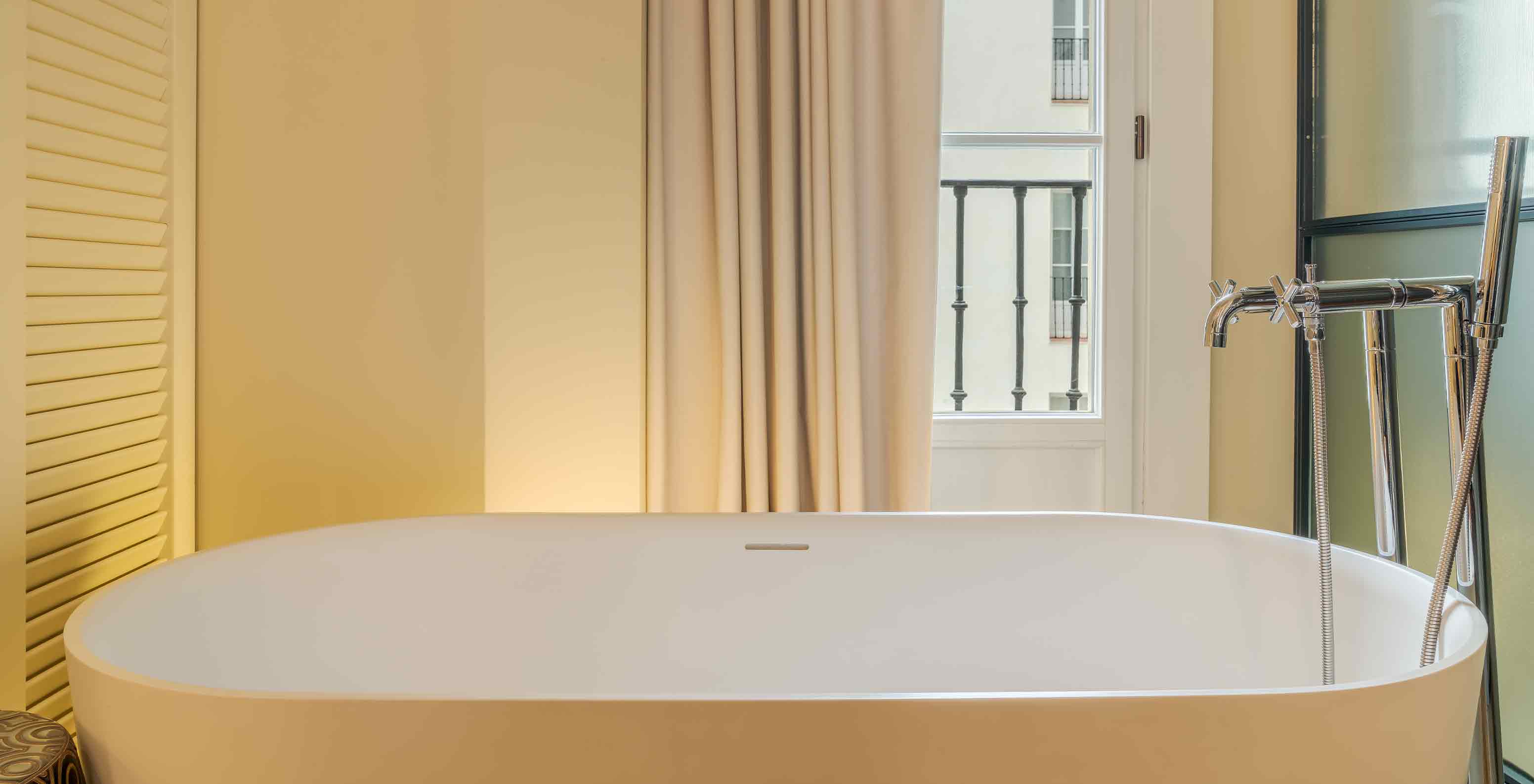 Suite at Pestana Collection Plaza Mayor with freestanding bathtub beside tall windows and beige curtains