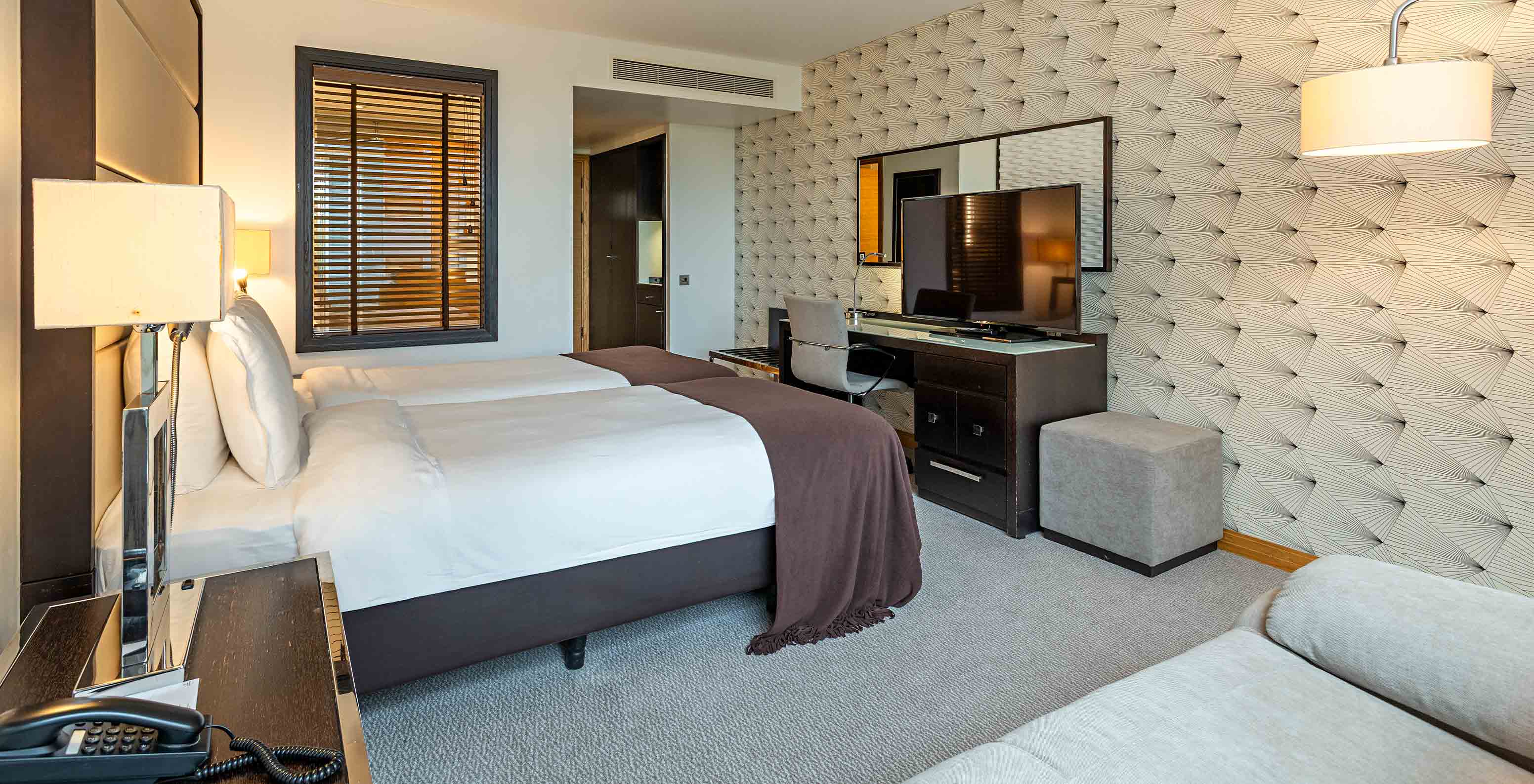 Modern hotel room with twin beds, patterned wall, desk with television and soft lighting beside the window