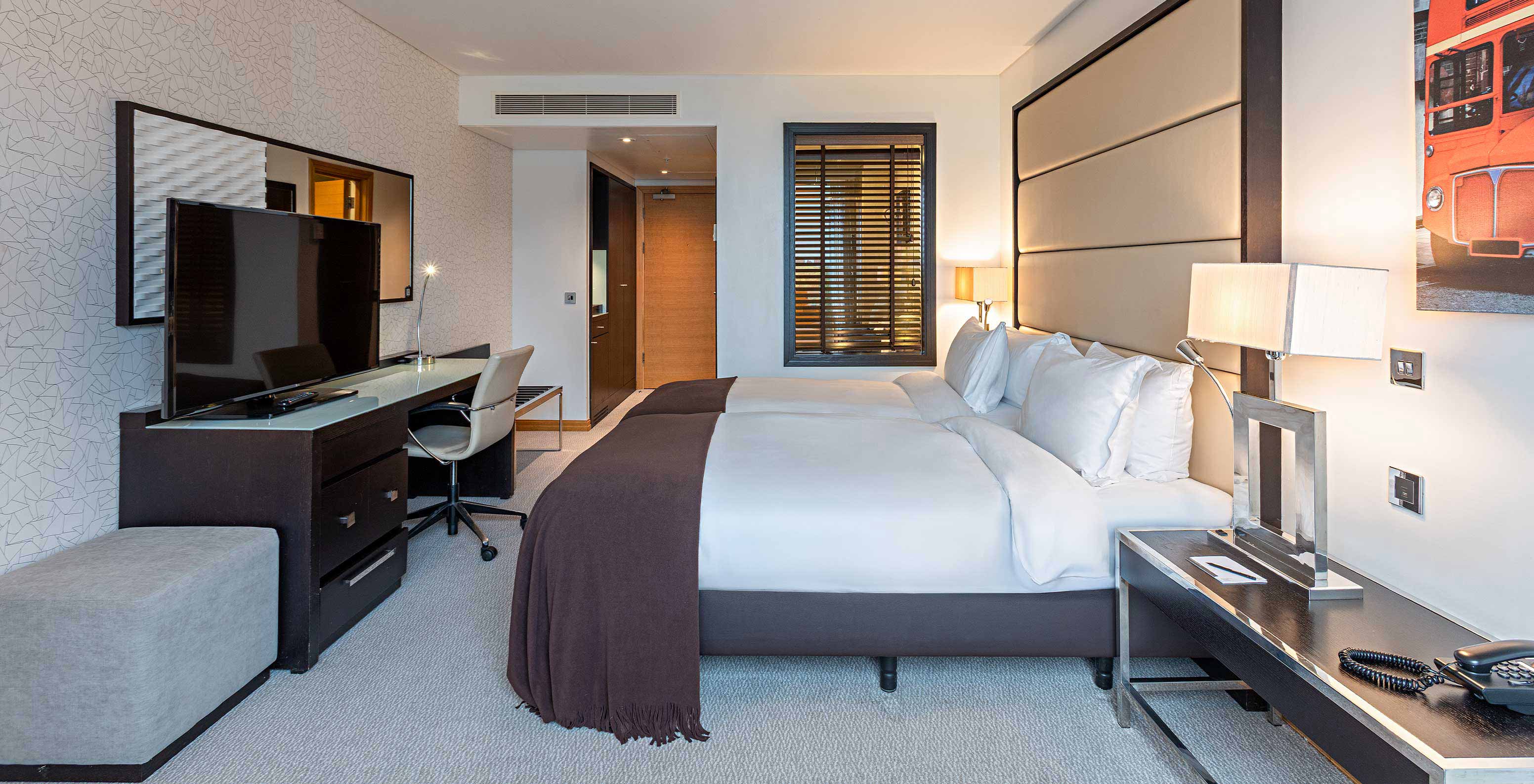 Modern hotel room with twin beds, desk, television and bedside lamps beside a window with dark shutters