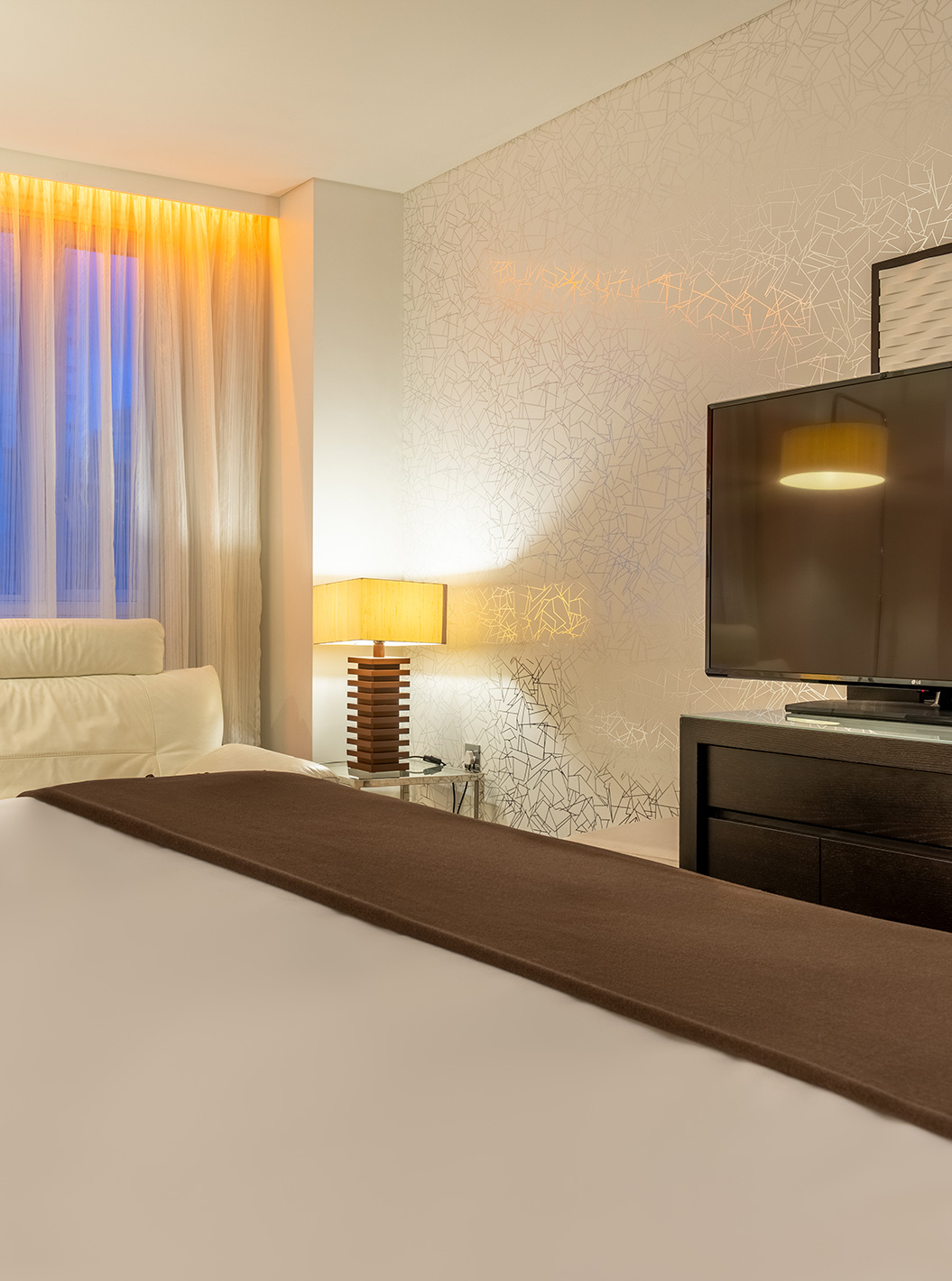 The Deluxe King at Pestana Chelsea Bridge has bed, desk with TV and chair, sofa and windows facing the city