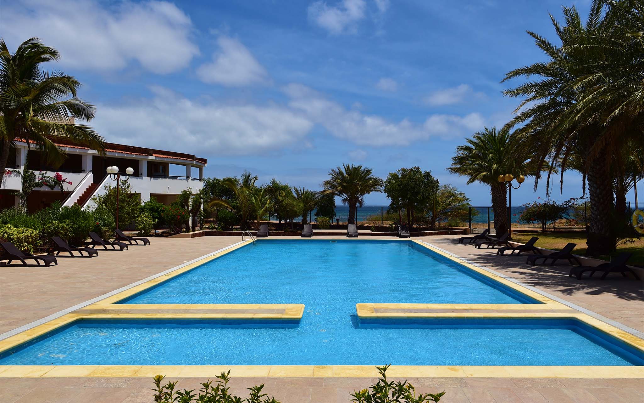 View of the outdoor pool at Pestana Tropico, with palm trees and sun loungers around the pool, and the sea in the background
