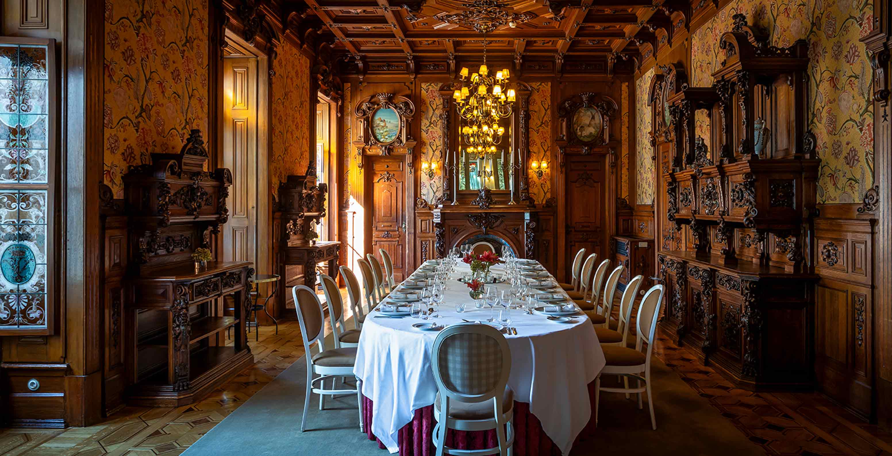 A formal dining room with a round table set for dinner, surrounded by ornate wood paneling and a chandelier.