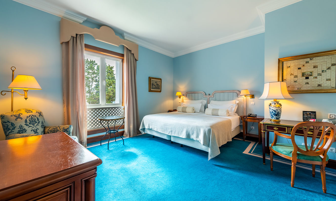 The Deluxe room at Pestana Palace Lisbon has two single beds, a window, an armchair, a desk, and a blue carpet