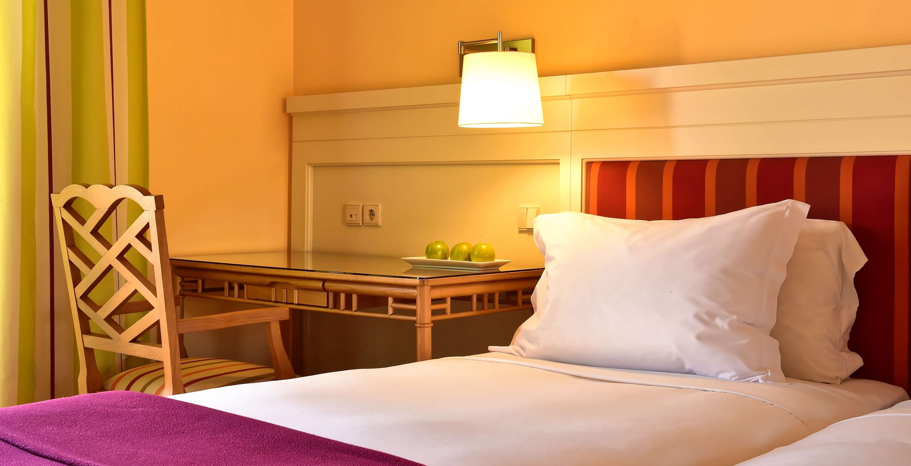 Warmly lit hotel room with white bedding, red headboard, wooden desk and chair beside a bedside lamp
