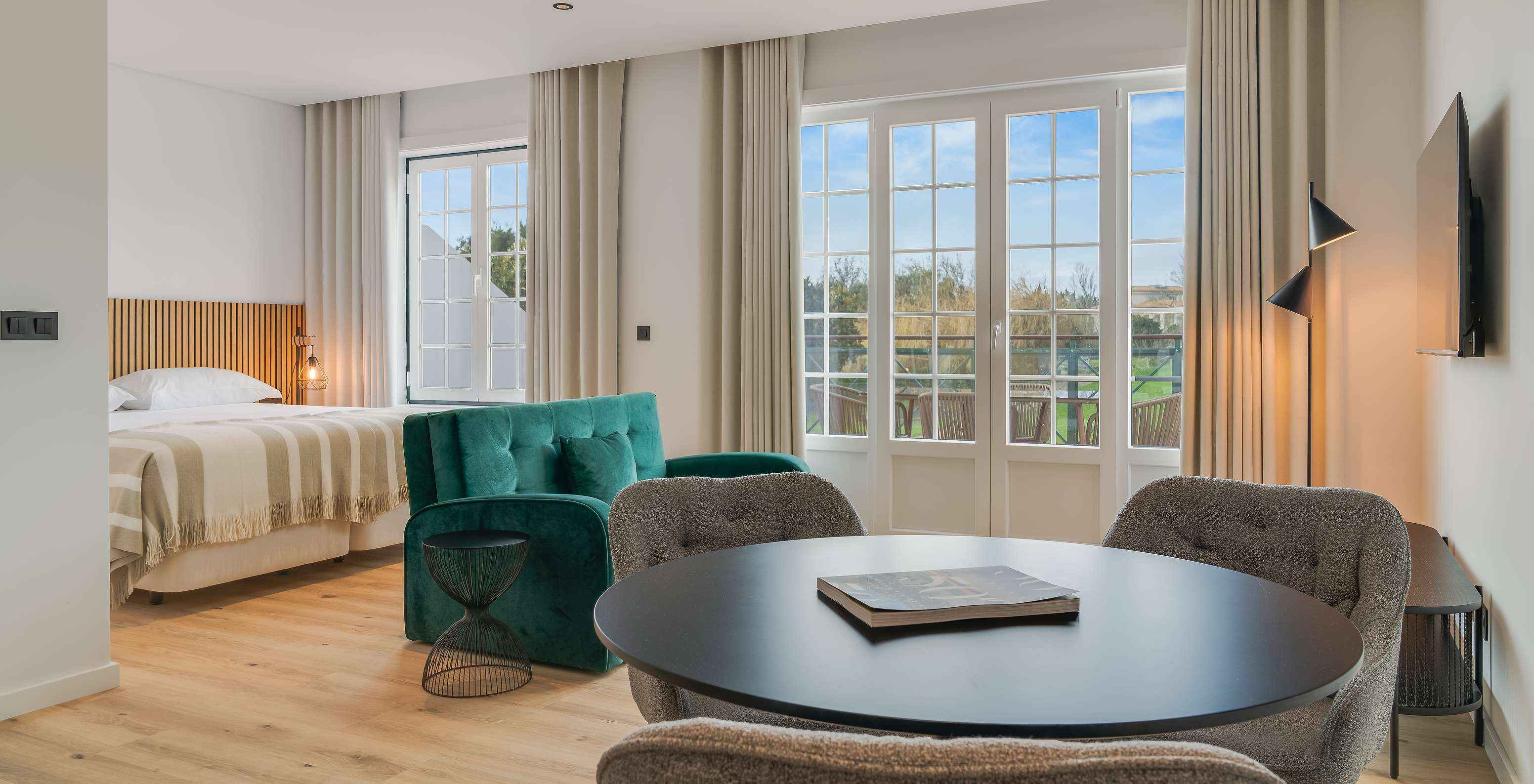 Bright hotel suite with bed, round dining table, green sofa and balcony doors overlooking trees outside
