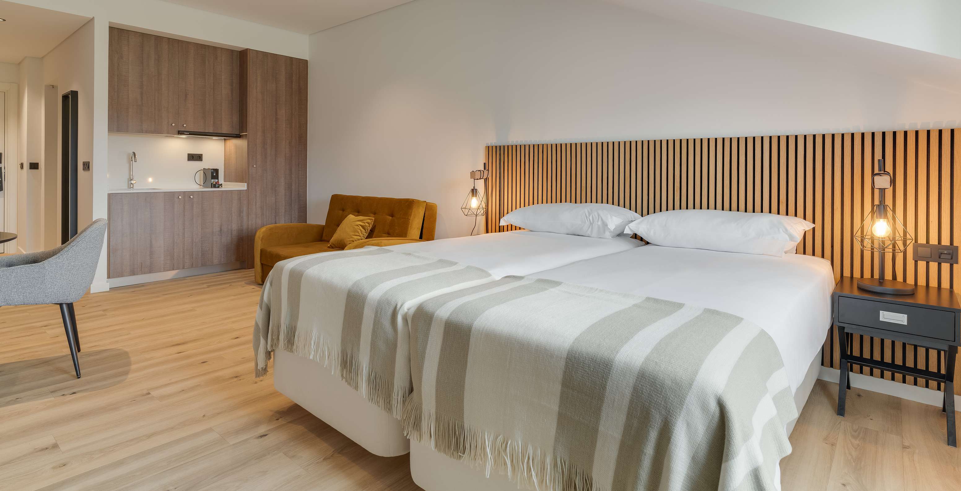Modern bedroom with twin beds, striped throws, wood panel headboard, lounge chair and compact kitchenette