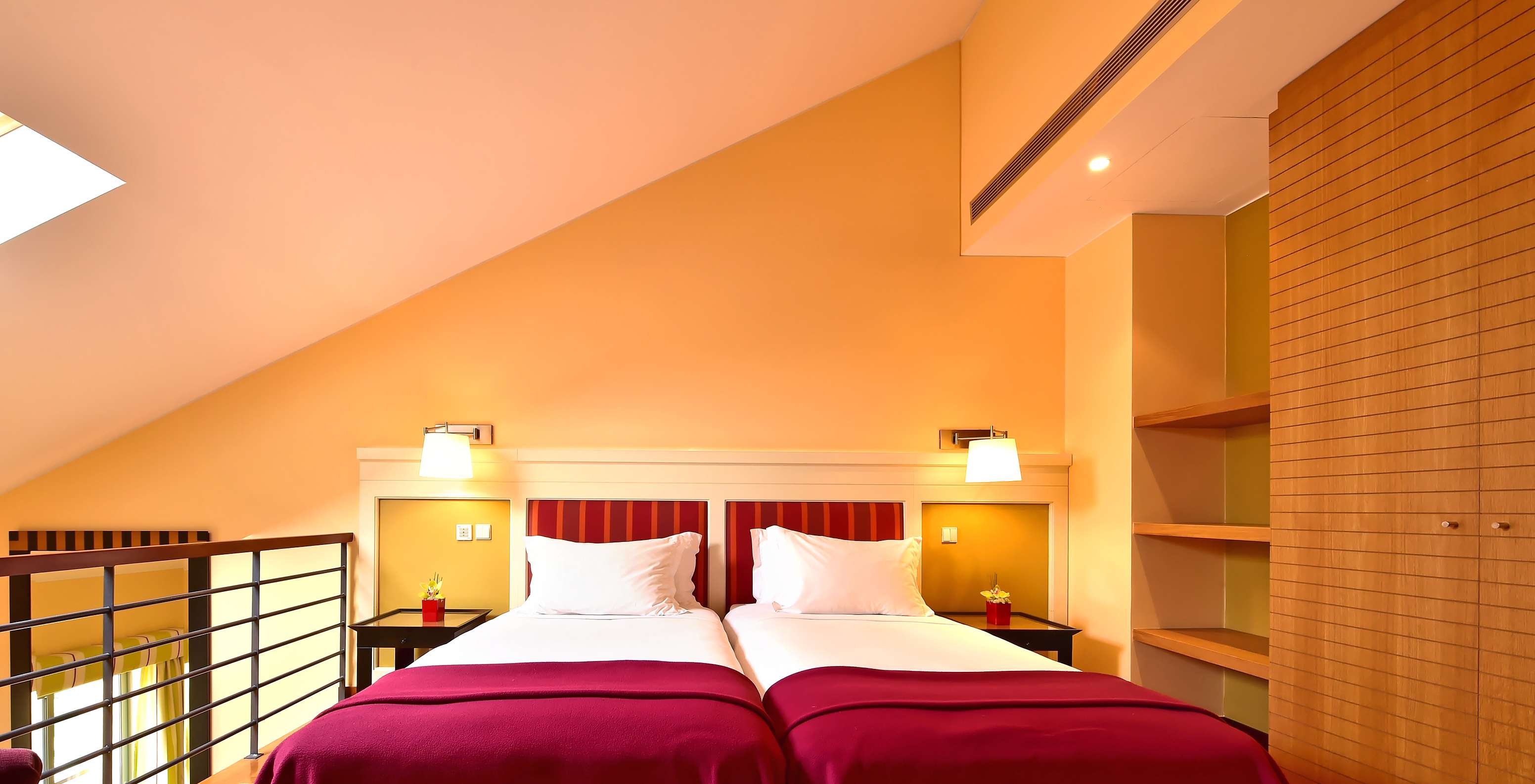 Warm attic bedroom with two single beds, bedside lamps, sloped ceiling and wooden shelving beside a mezzanine