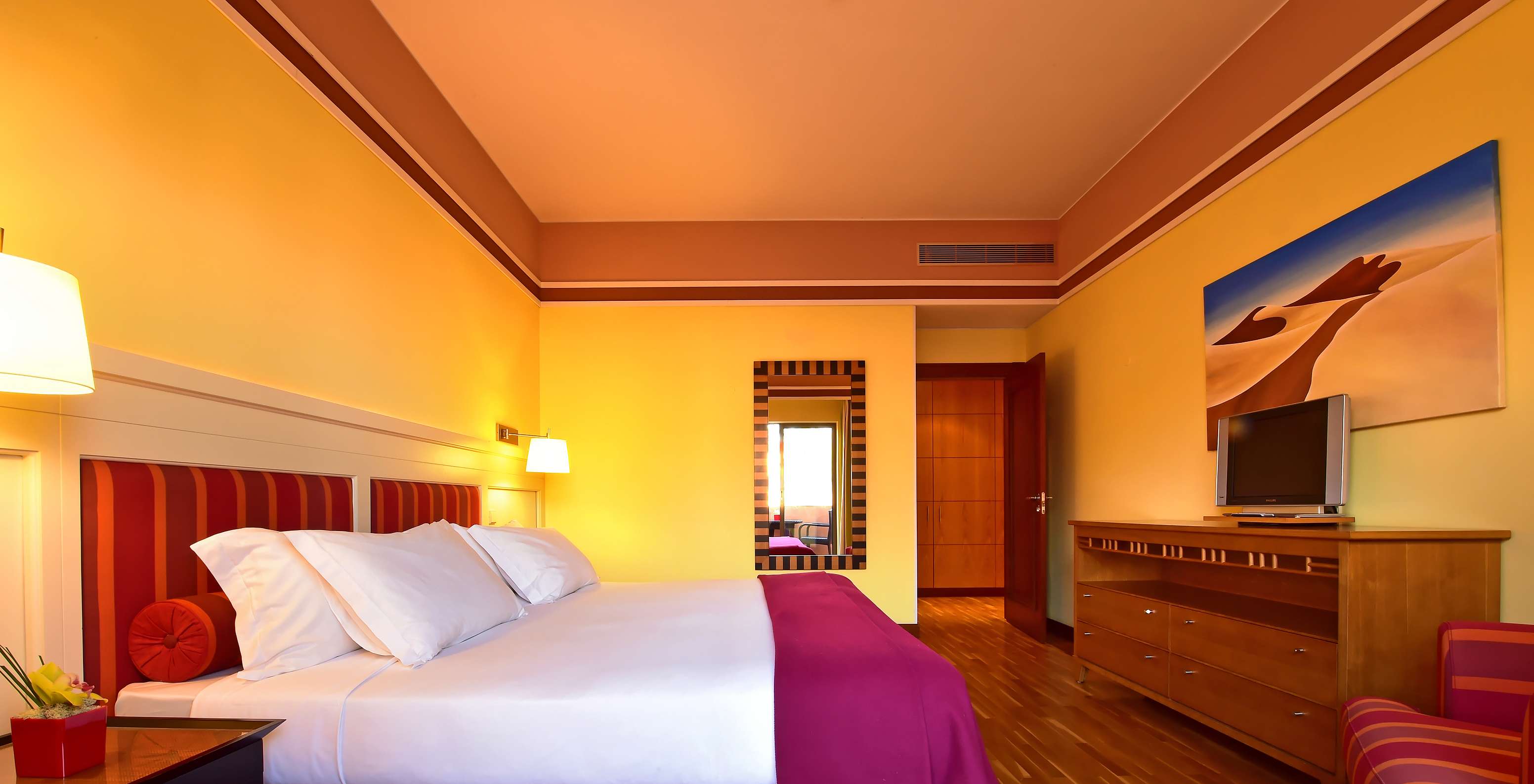 Warm hotel bedroom with striped headboard, wooden dresser, wall mirror and television beside the bed