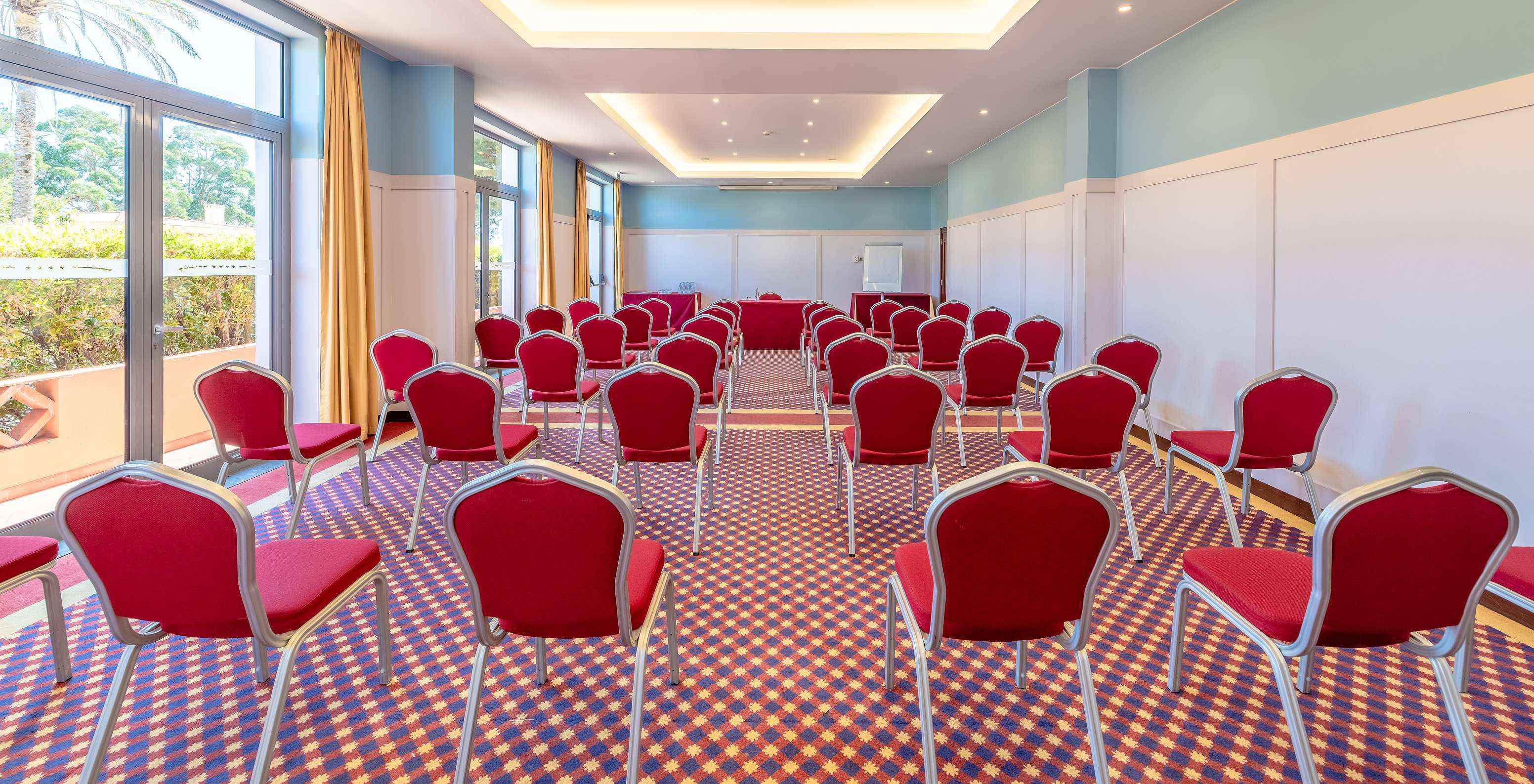 Bright meeting room with red chairs in rows, patterned carpet, large windows and recessed ceiling lights