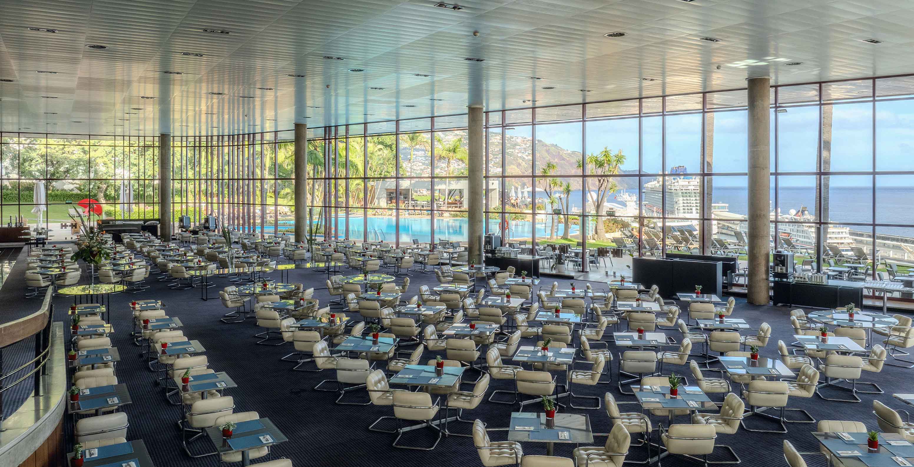 Panoramic view of a restaurant with several tables, a ramp and a large glass overlooking the pool and the sea