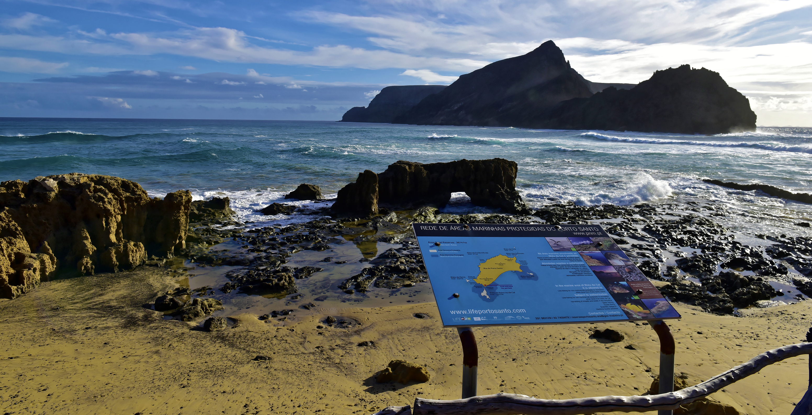 Calheta beach in Porto Santo with an informative map and features rocks, waves, blue sky, and white clouds