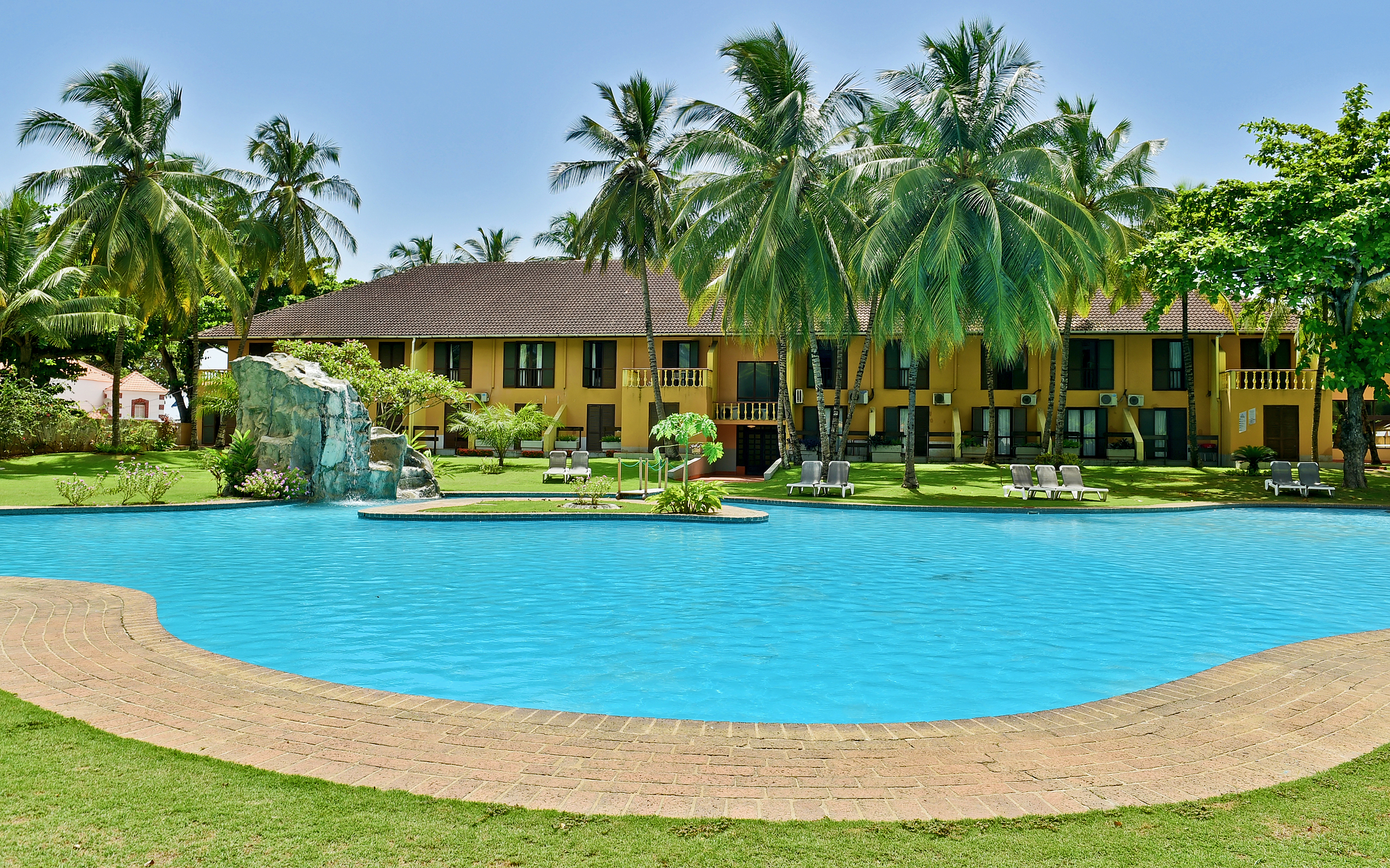 View of the yellow building with several windows, outdoor pool, palm trees, and sun loungers of a 4-star hotel in São Tomé