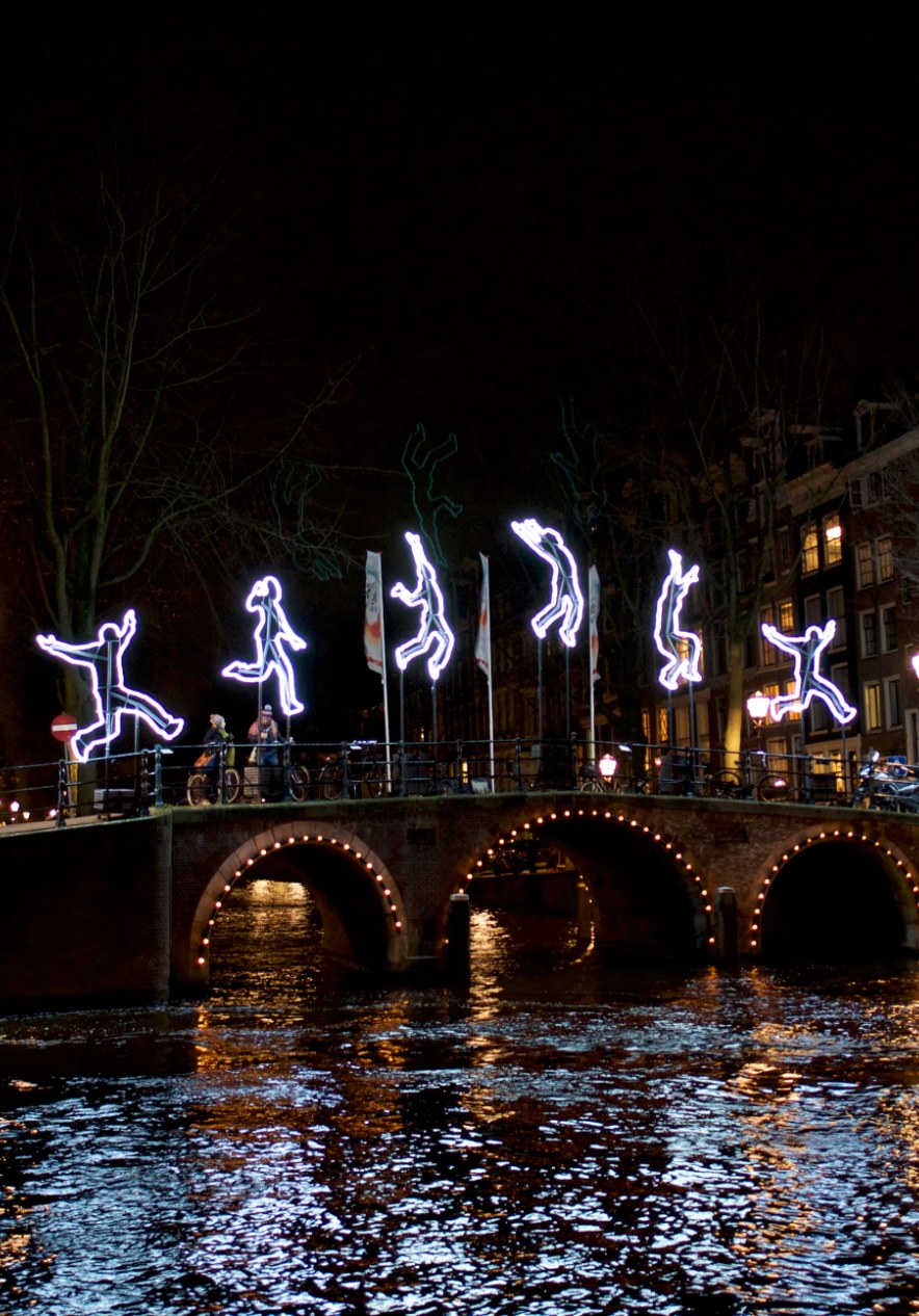 Light figure installations above a canal bridge in Amsterdam at night with reflections shimmering on the water