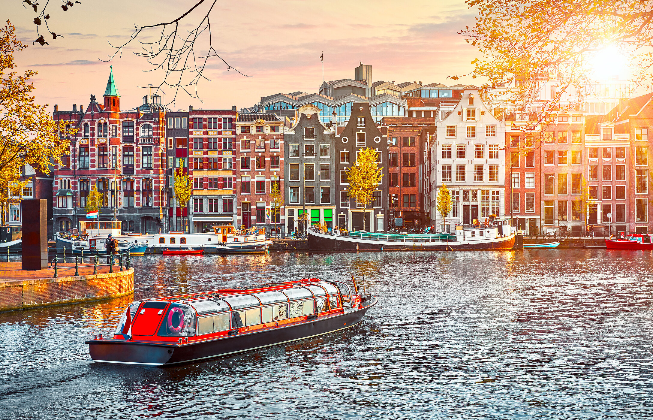 Picturesque view of the canals of Amsterdam, with typical Dutch houses, a red tourist boat, and a sky with warm tones