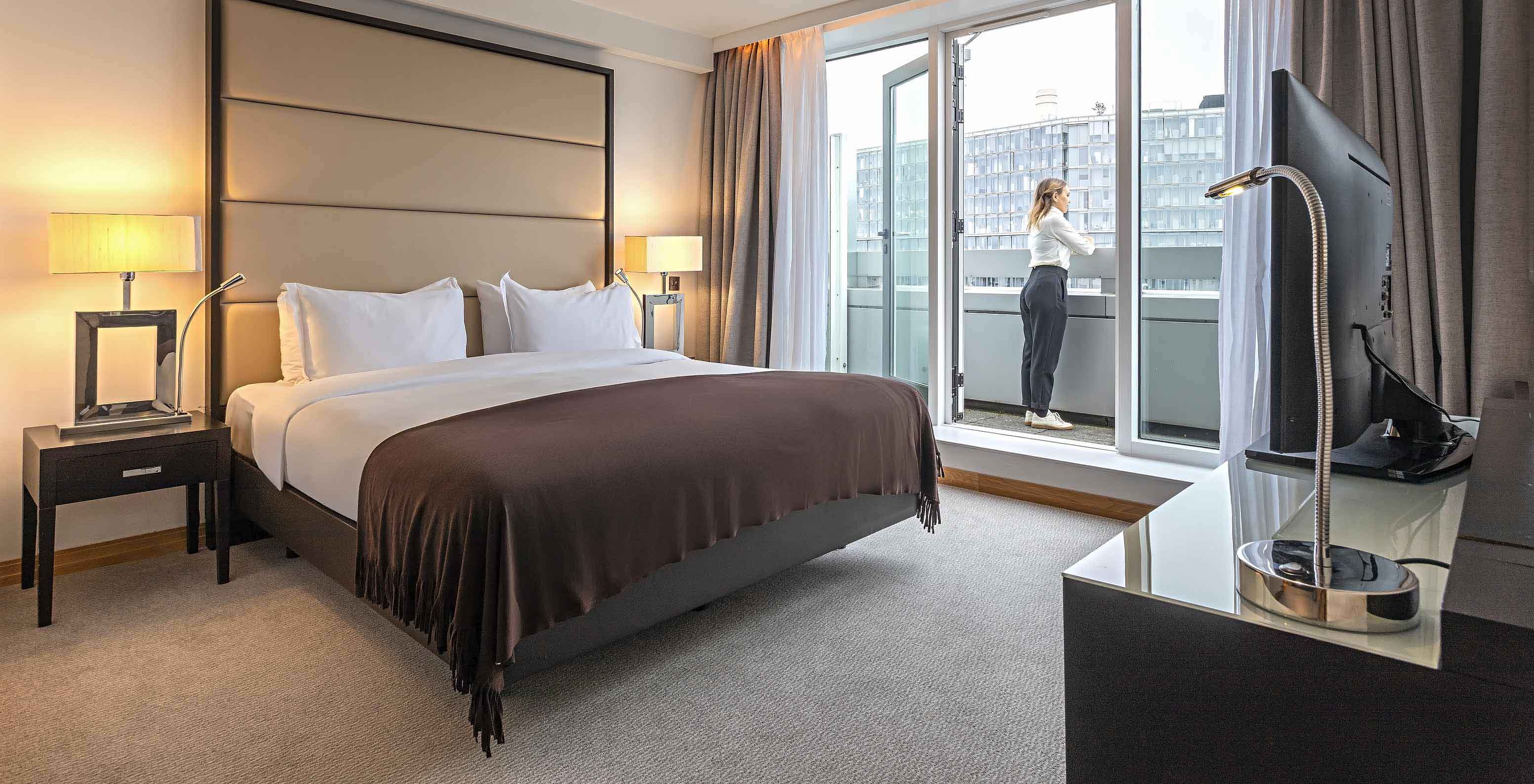 Modern hotel bedroom with large bed, desk and open balcony doors as a guest overlooks nearby buildings