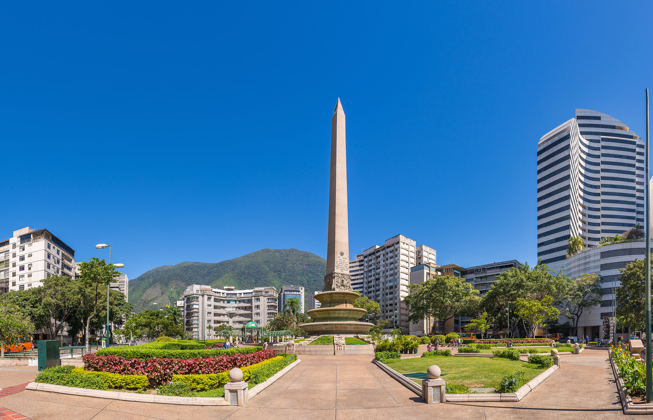 Obelisk Monument, a large stone column in the middle of a park with flowers, surrounded by tall buildings in Venezuela.