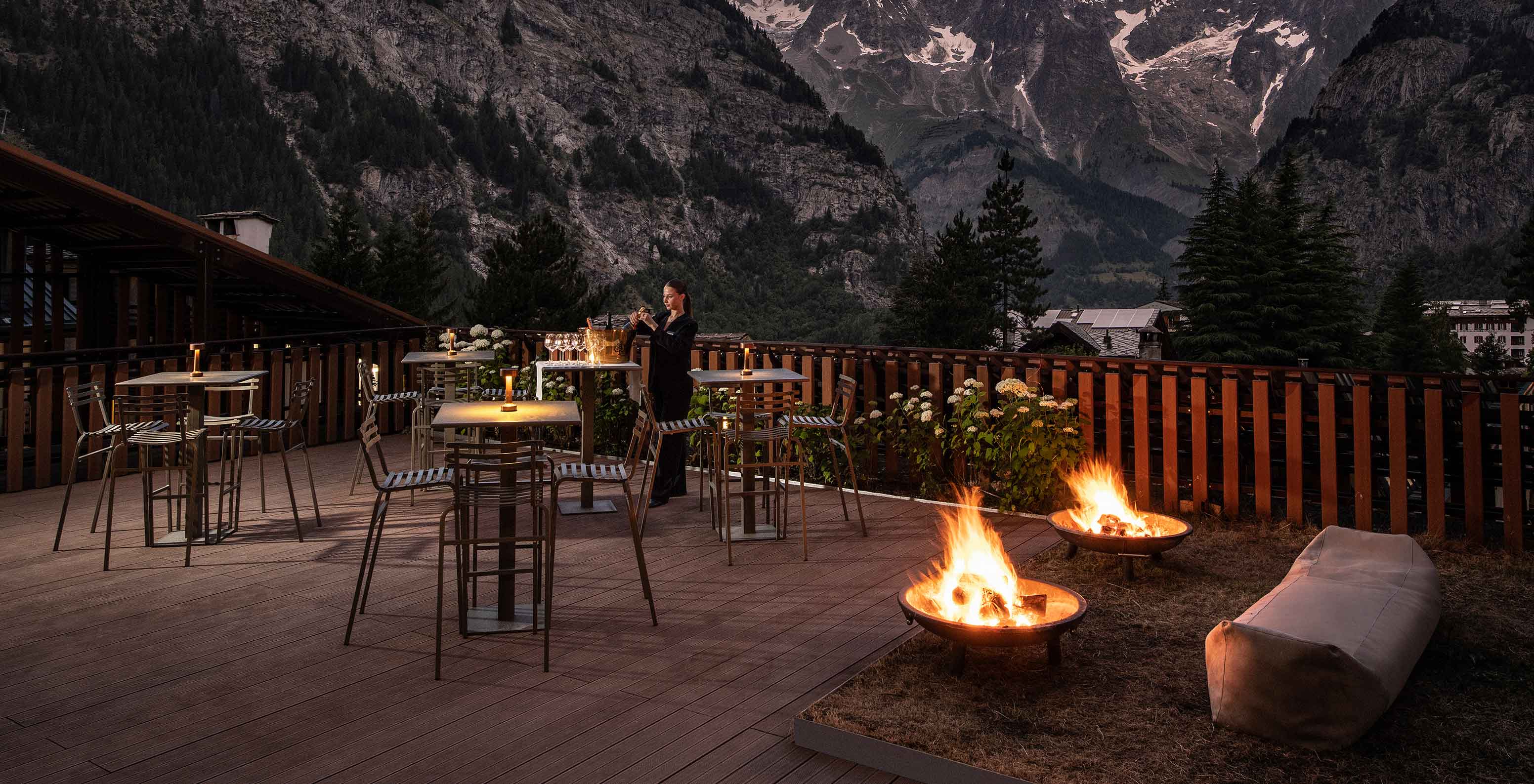 Terraza de montaña al atardecer con mesas altas iluminadas, fogones encendidos y picos nevados al fondo