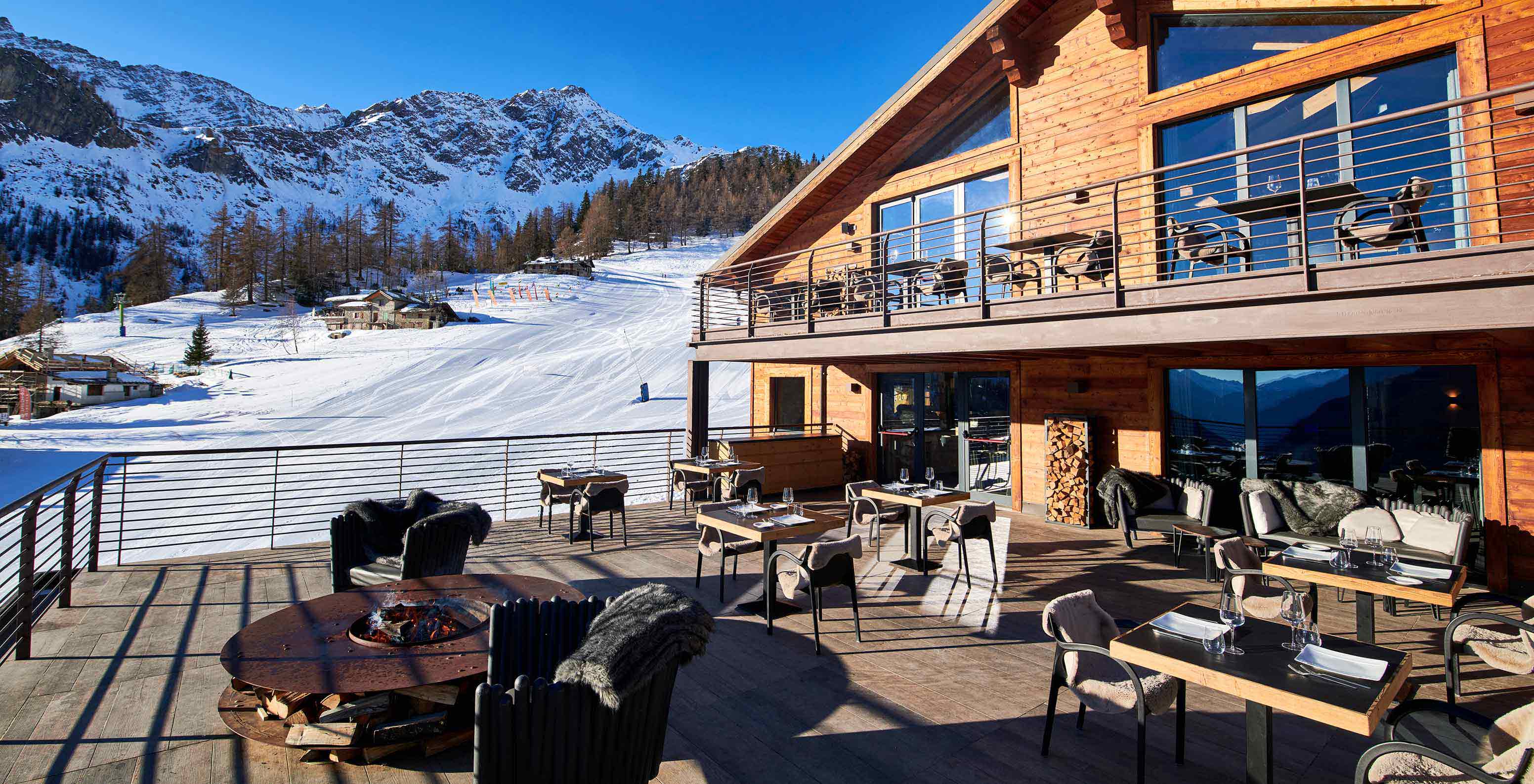 Terraza de madera con mesas y sillones frente a pistas nevadas y montañas bajo un cielo azul despejado
