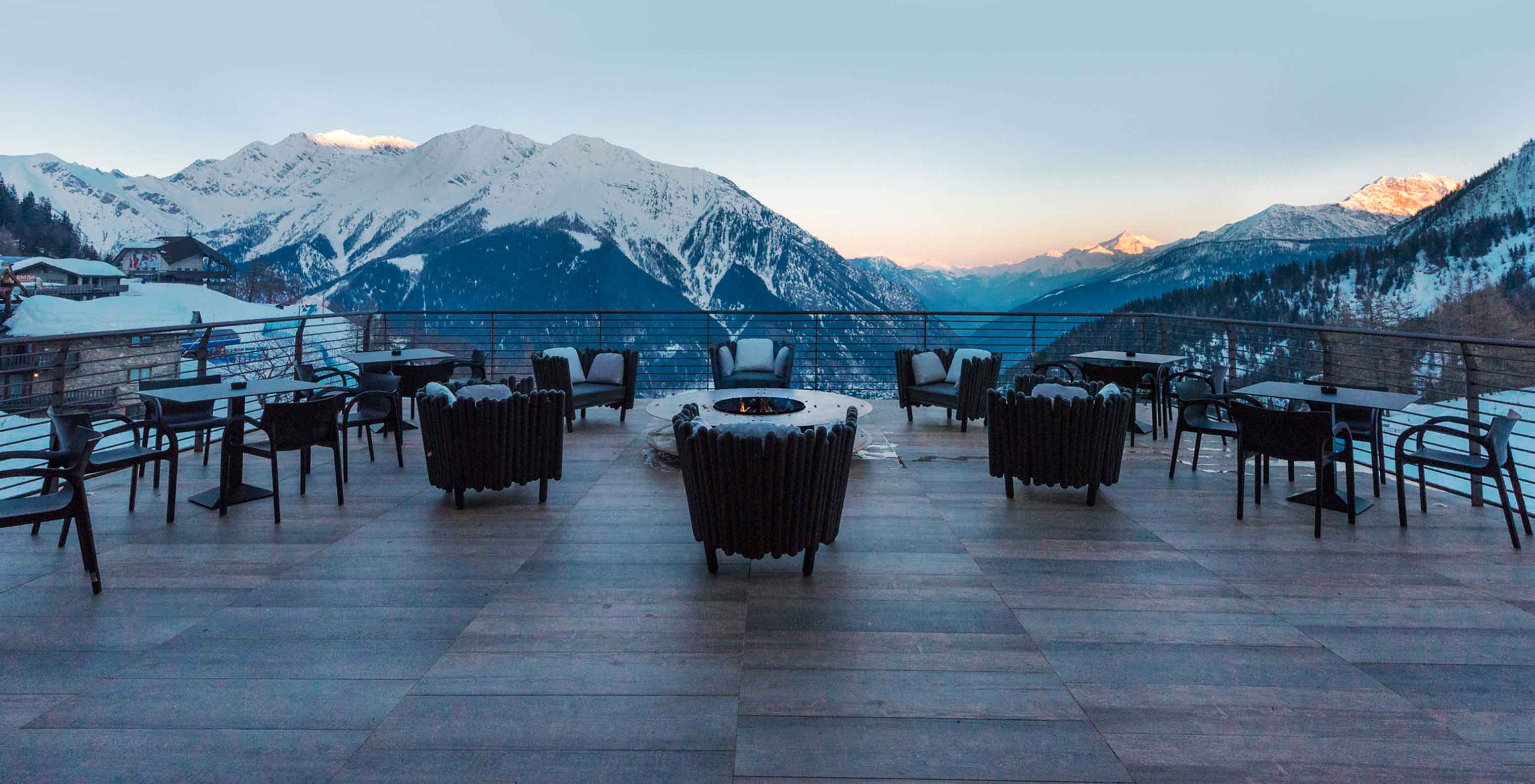 Terraza con mesas y sillones frente a montañas nevadas, con fogón central y vista amplia del valle alpino
