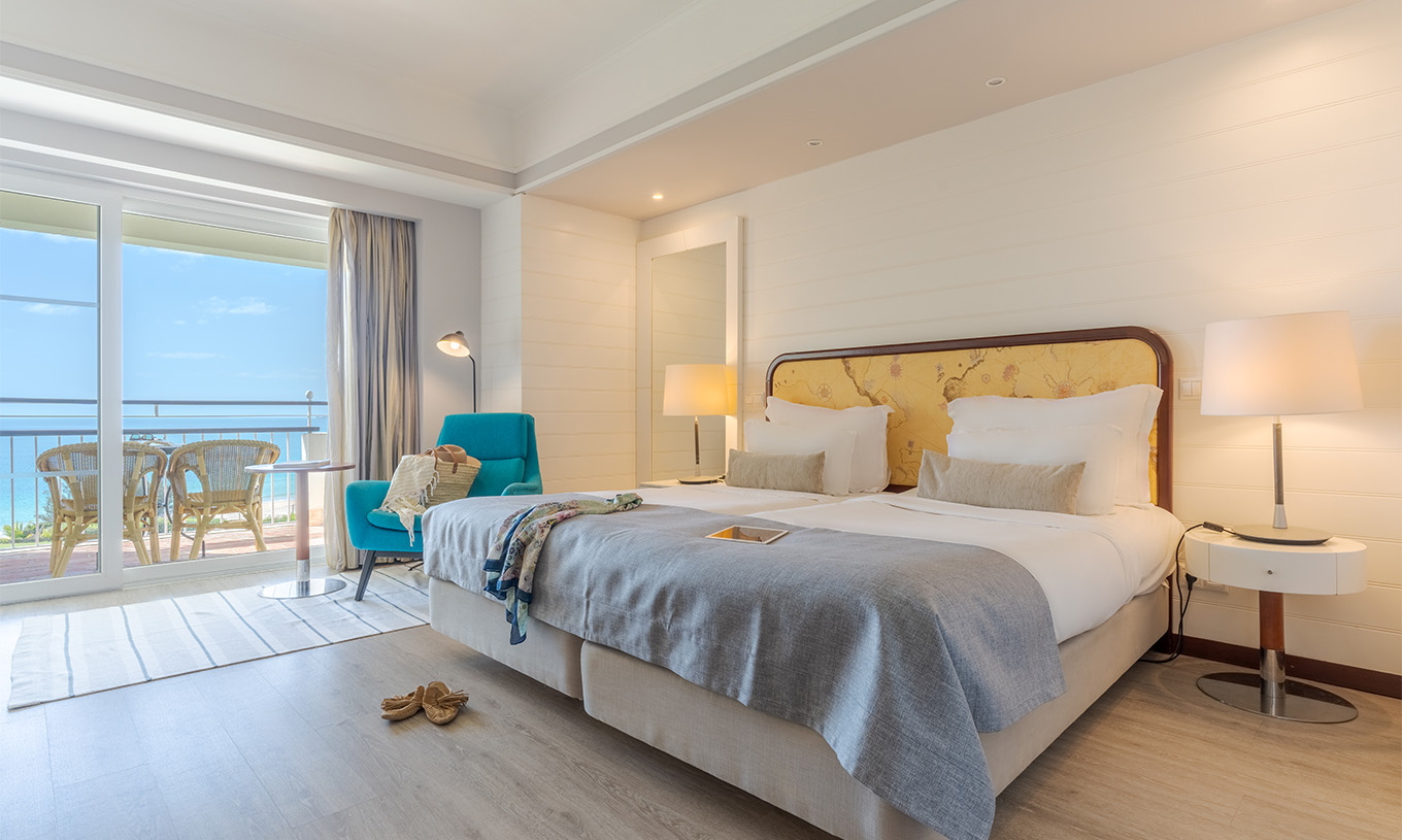 El Deluxe Vista Mar del Pestana Alvor Praia tiene dos camas juntas con sillón azul, mesa de apoyo y terraza con vista al mar
