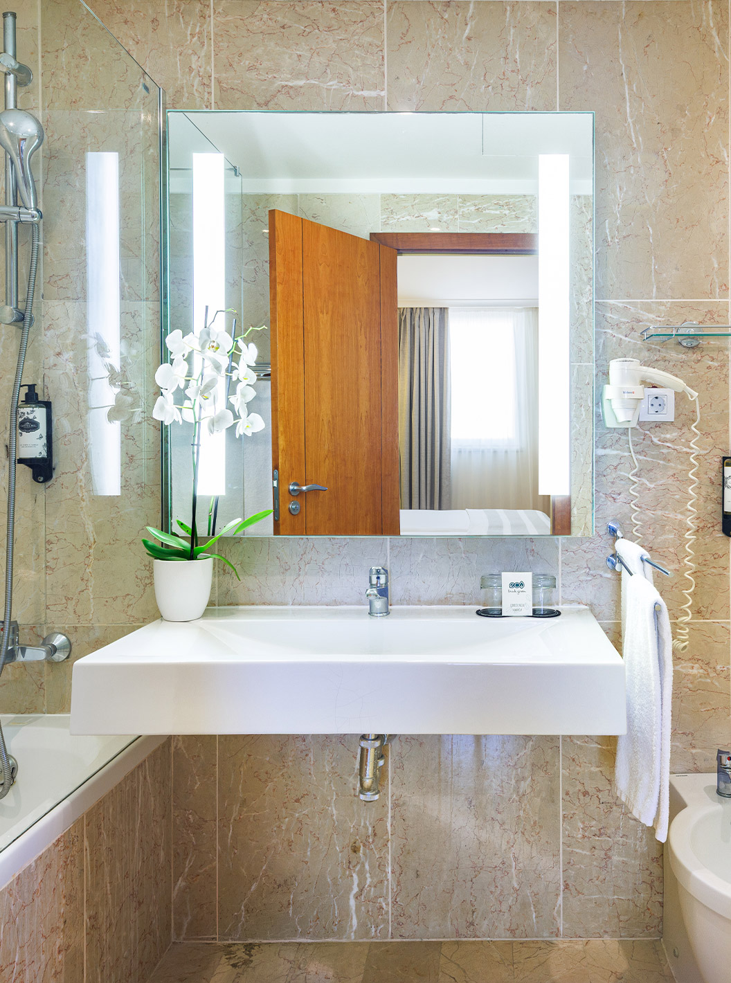 Suite del Pestana Viking con baño de mármol beige, espejo iluminado, ducha y lavabo moderno con orquídea blanca