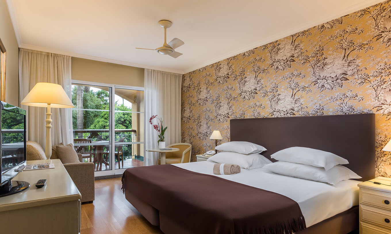 El Studio Classic del Pestana Village tiene una cama de casal con cabecero y colcha marrón, papel tapiz floral y balcón
