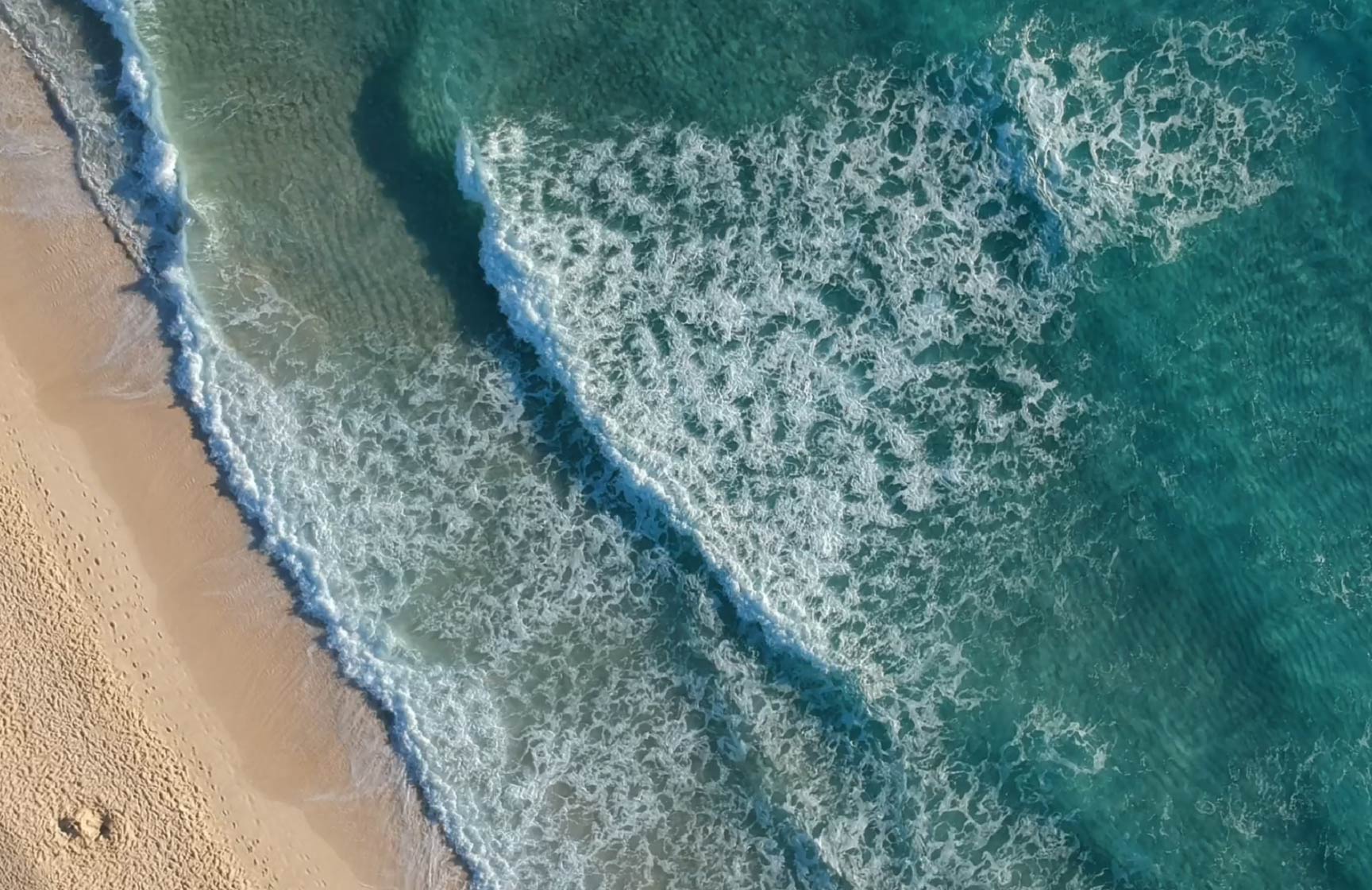Vista aérea de una playa con olas rompiendo en la arena dorada, en un hotel del Pestana Hotels & Resorts