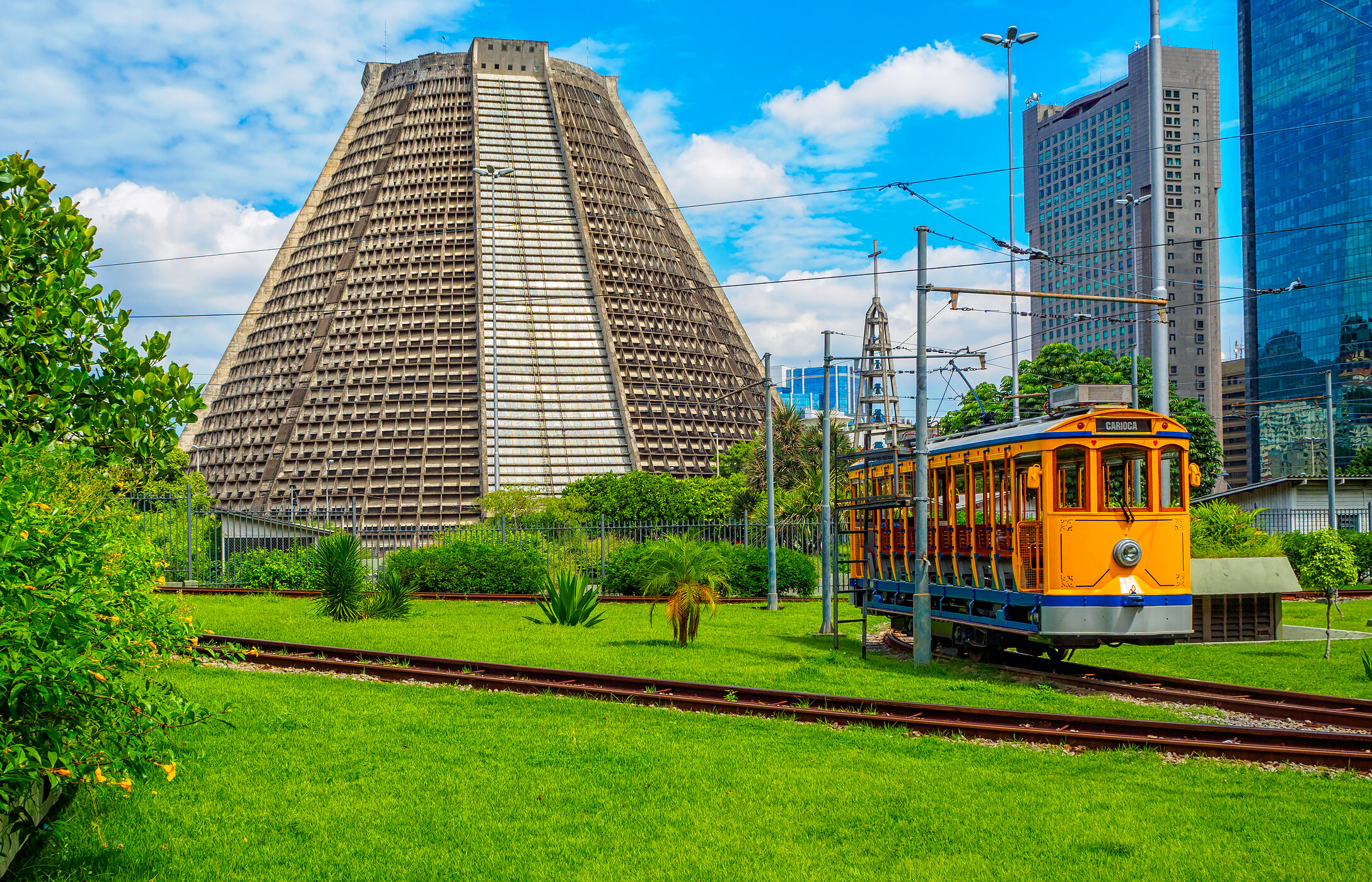 Catedral Metropolitana de Río de Janeiro, rodeada de vegetación, un tranvía antiguo y edificios al fondo.