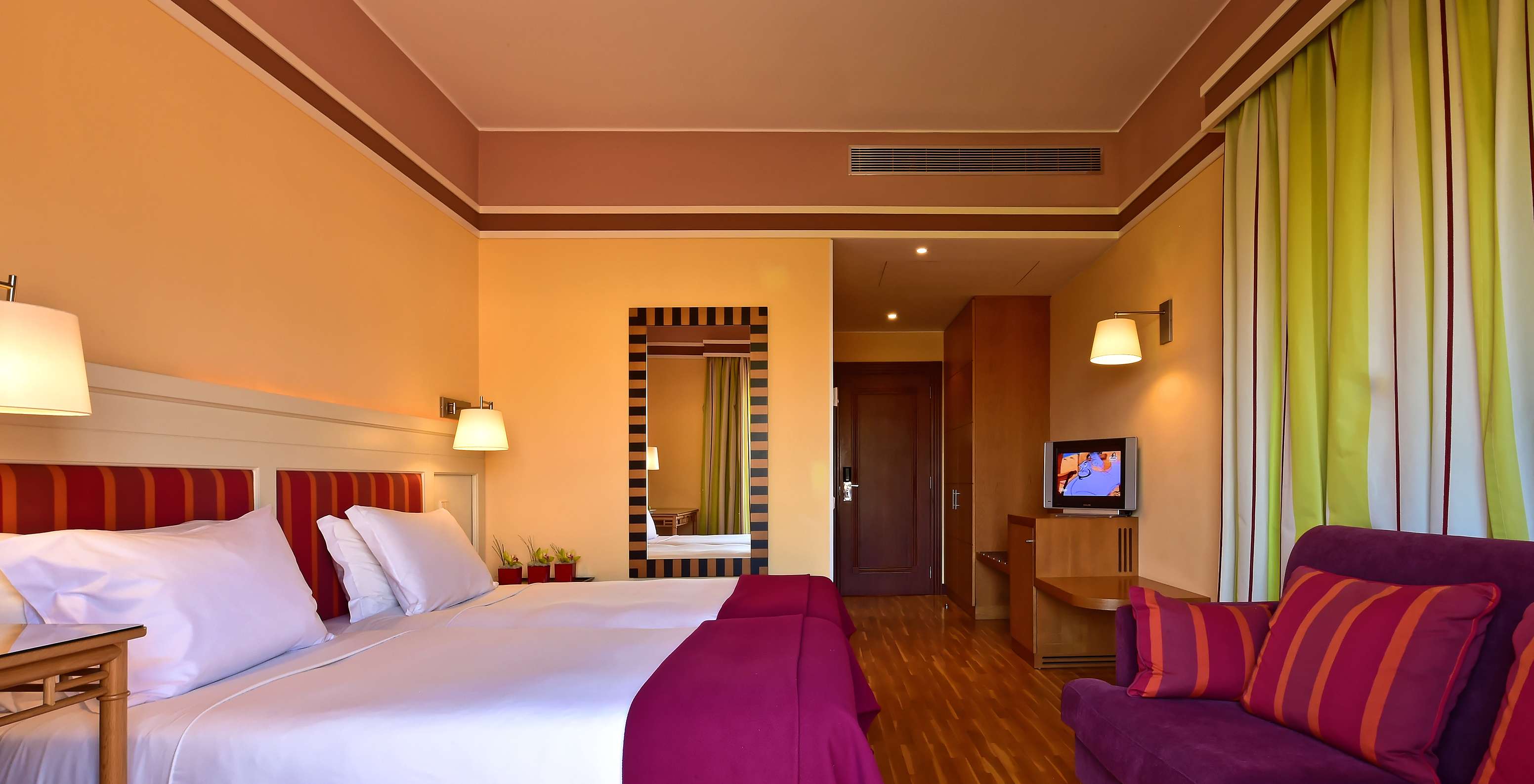 Habitación con dos camas, sofá morado, escritorio y cortinas verdes junto a ventana amplia e iluminación cálida