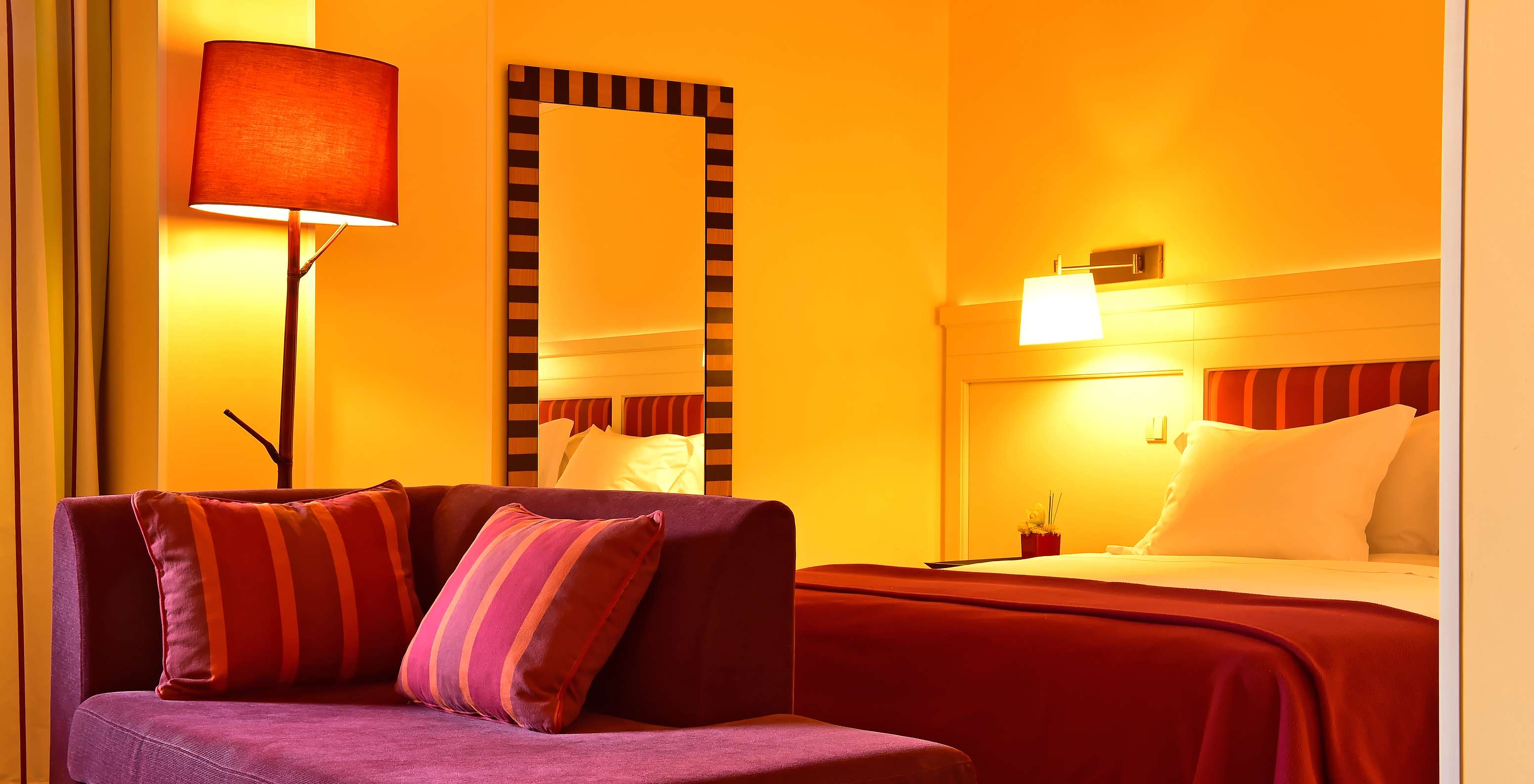 Habitación iluminada con sofá morado, cama con cabecero rojo, espejo alto y lámparas de luz cálida tenue