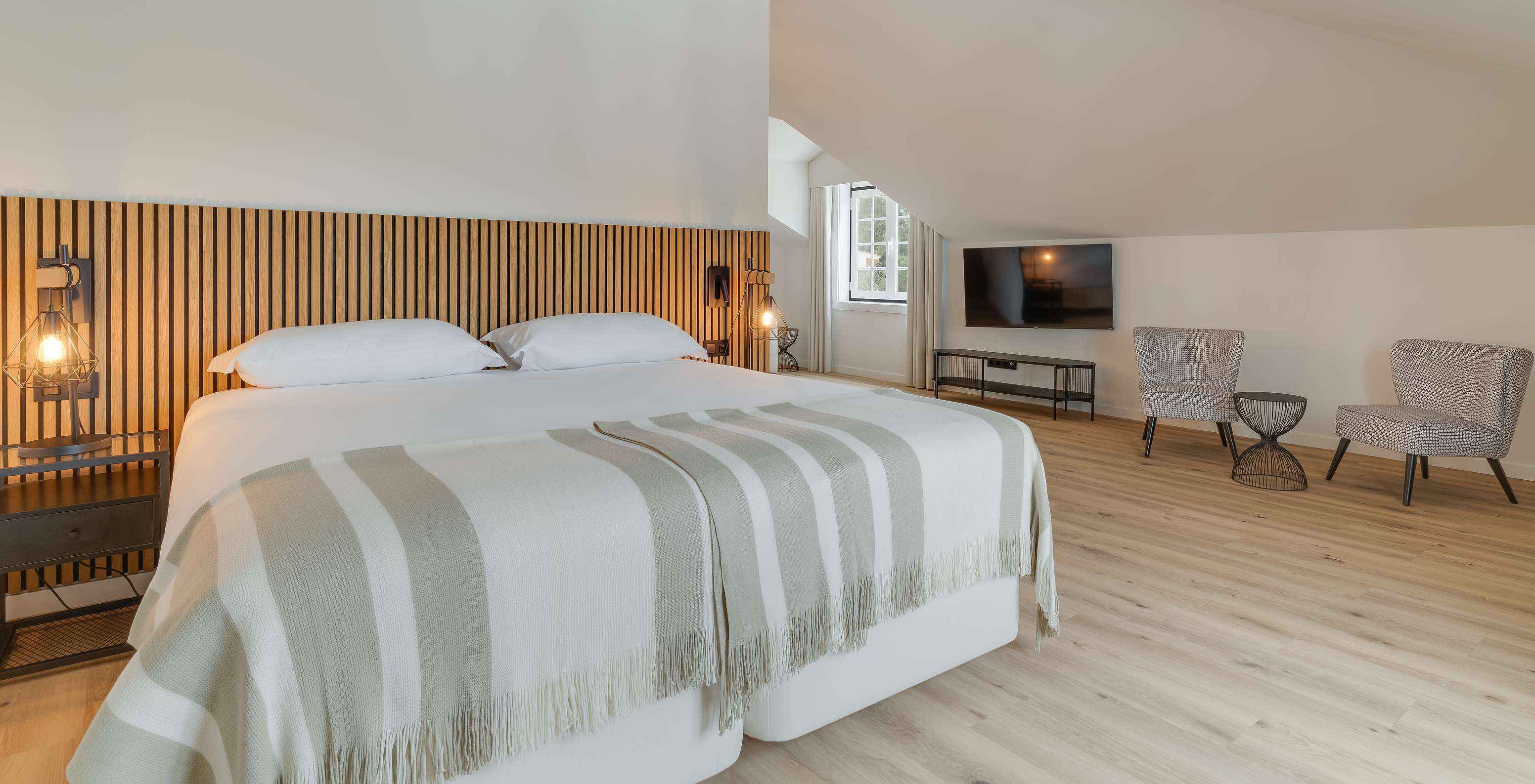 Dormitorio amplio con cama doble, cabecero de madera, sillones junto a la ventana y televisor mural