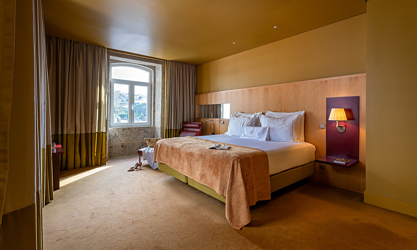 La Junior Suite tiene una cama doble con una manta beige y una ventana de piedra con vista al río